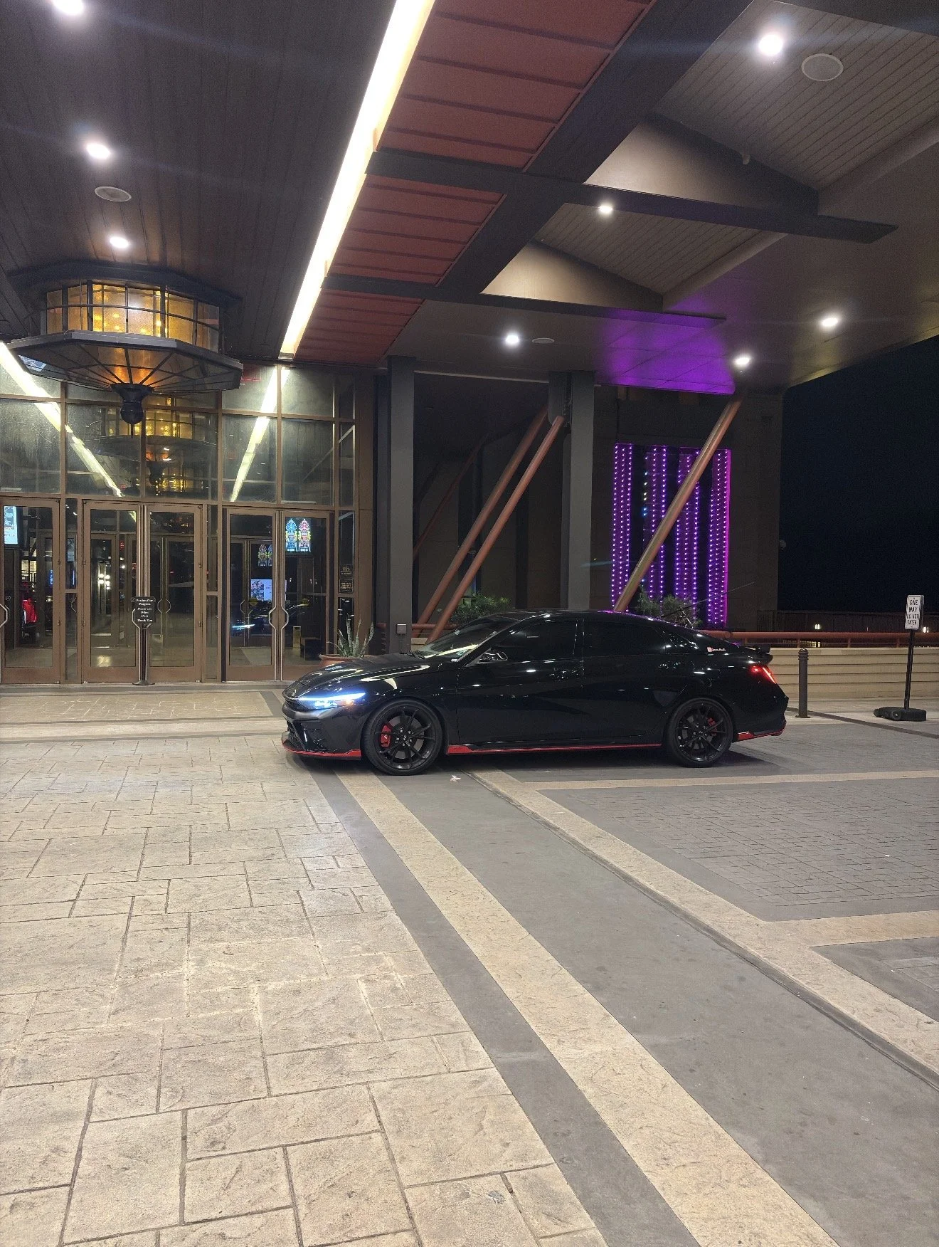 A black sports car parked in front of a modern building at night, with illuminated purple and yellow lighting and glass doors.