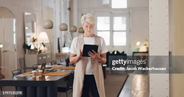 Older woman with white hair looking at a tablet in a bright modern office or co-working space.