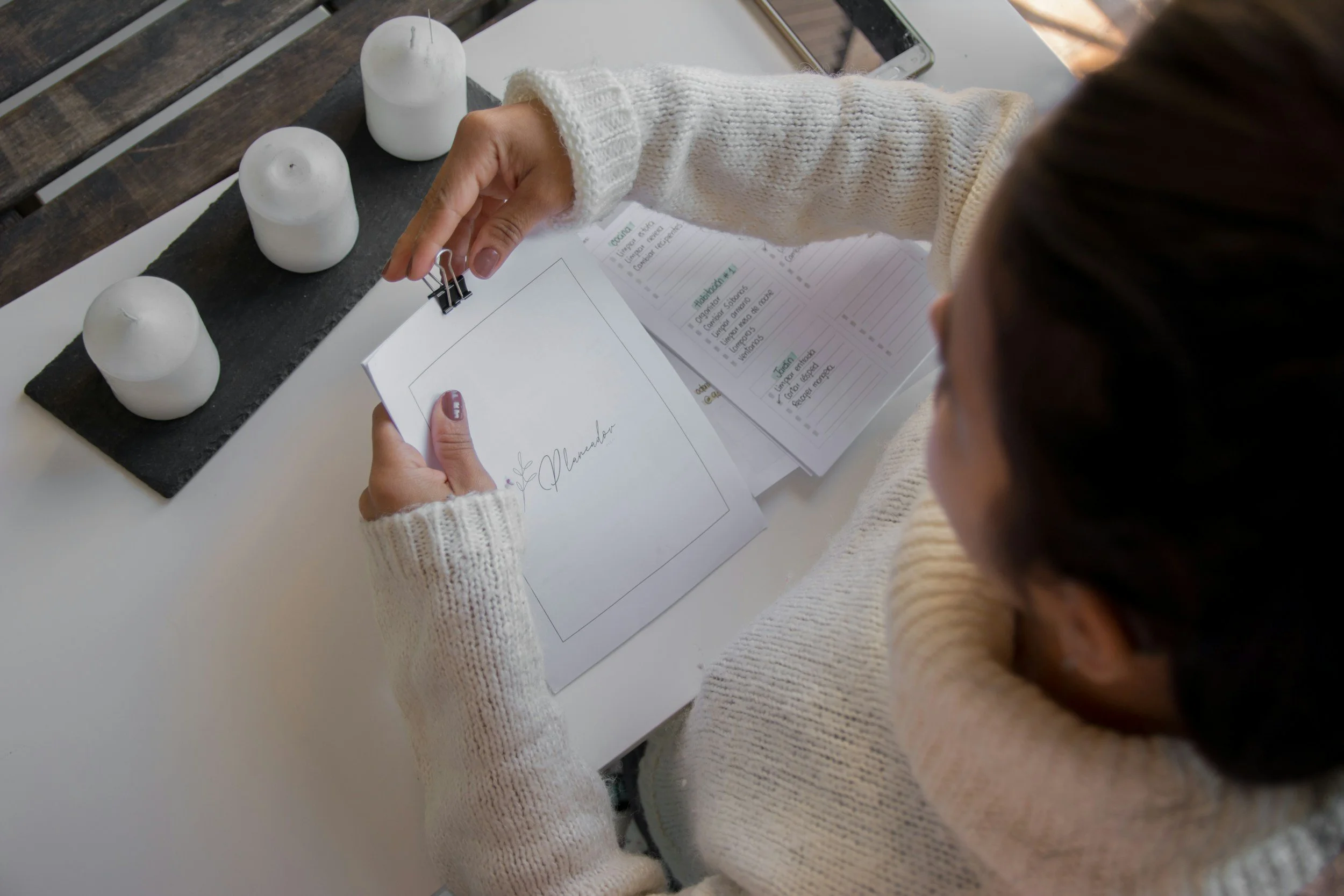 Person in cream-colored sweater organizing papers with white candles, a smartphone, and a clipboard on a table.