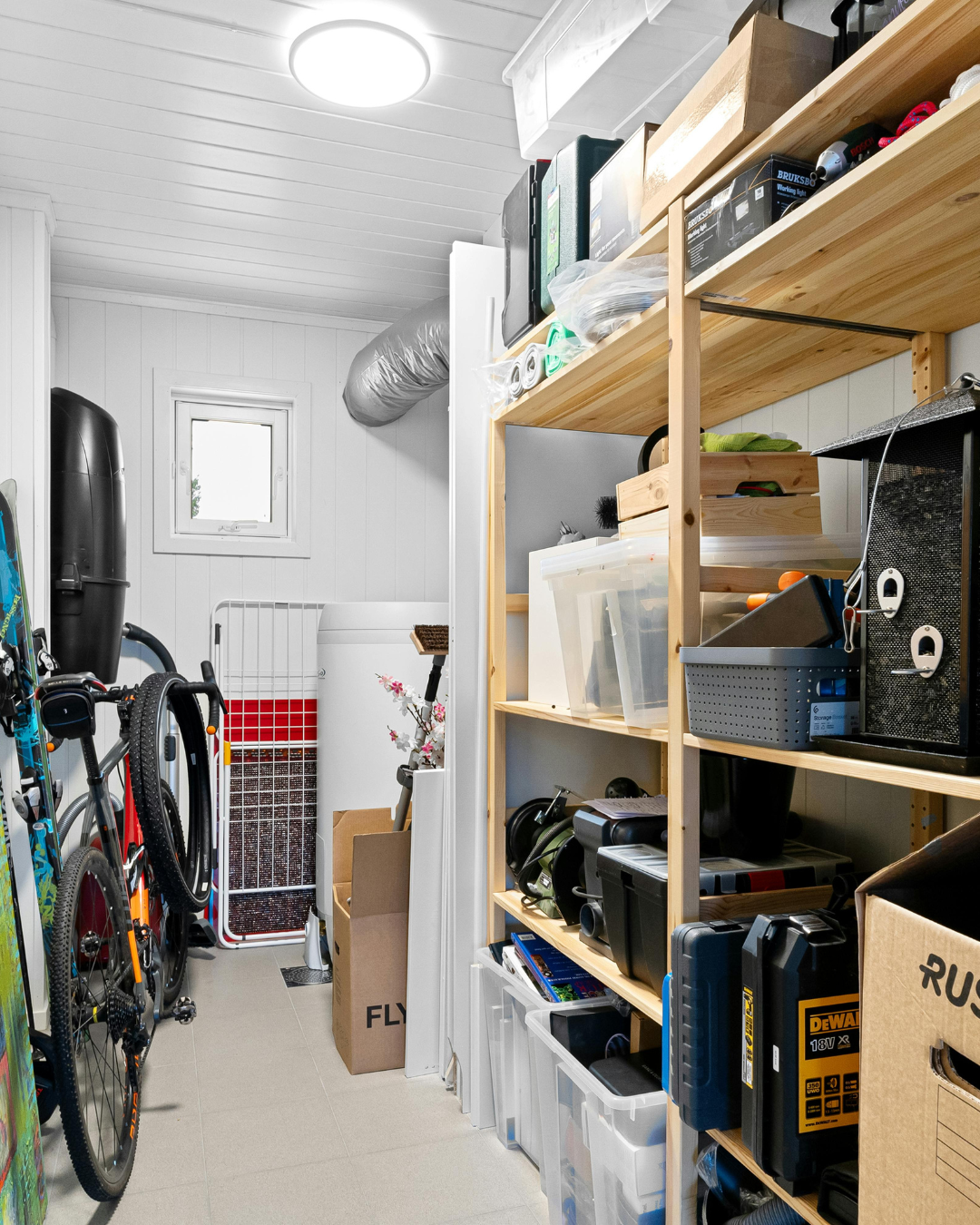 A storage room with white paneled walls and ceiling, containing bicycles, cardboard boxes, plastic storage bins, and various tools and equipment on wooden shelves.