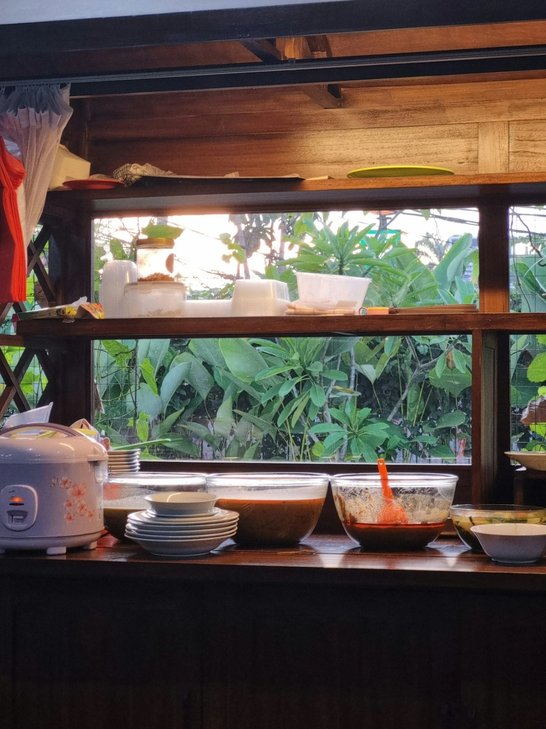 A lived-in home workspace with wooden shelving, storage containers, and greenery outside the window.