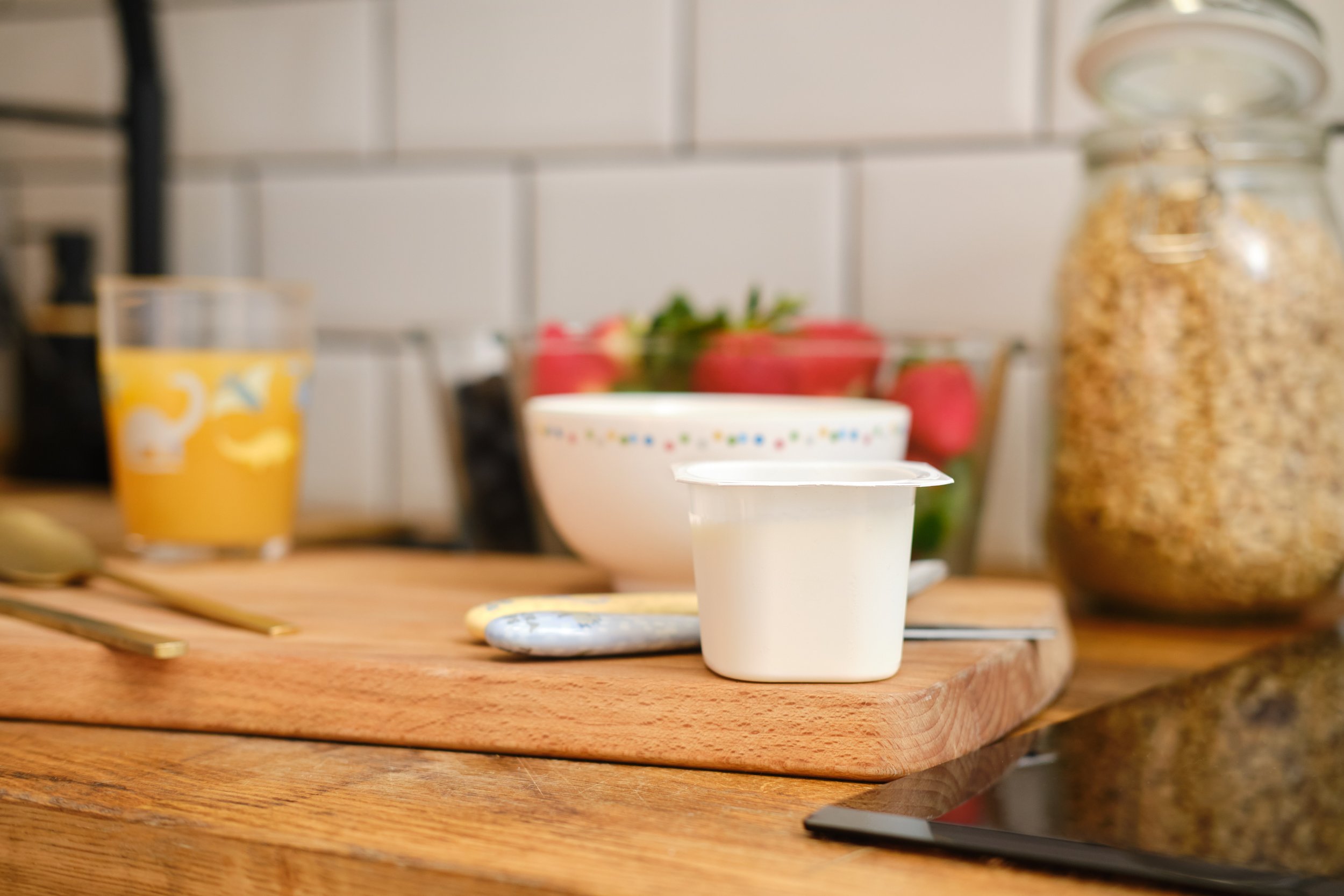 Close-up of a wooden kitchen counter with a small white bowl in the center and a white cup beside it. In the background, there are red strawberries, a cereal jar, and a glass of orange juice.