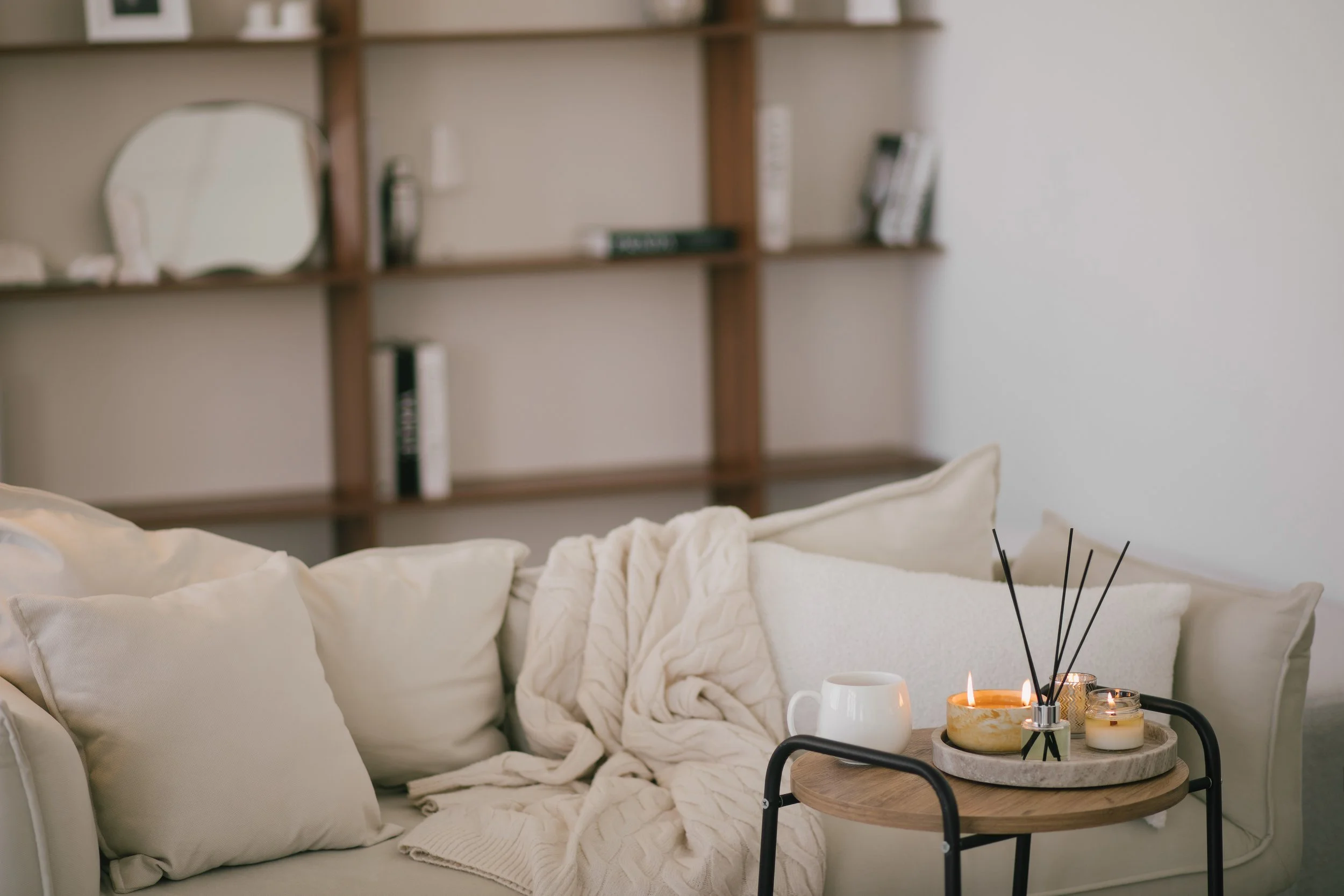 Cozy living room with white cushions and blanket on a sofa, a round wooden table with candles and reed diffusers, and a wooden bookshelf on the wall.