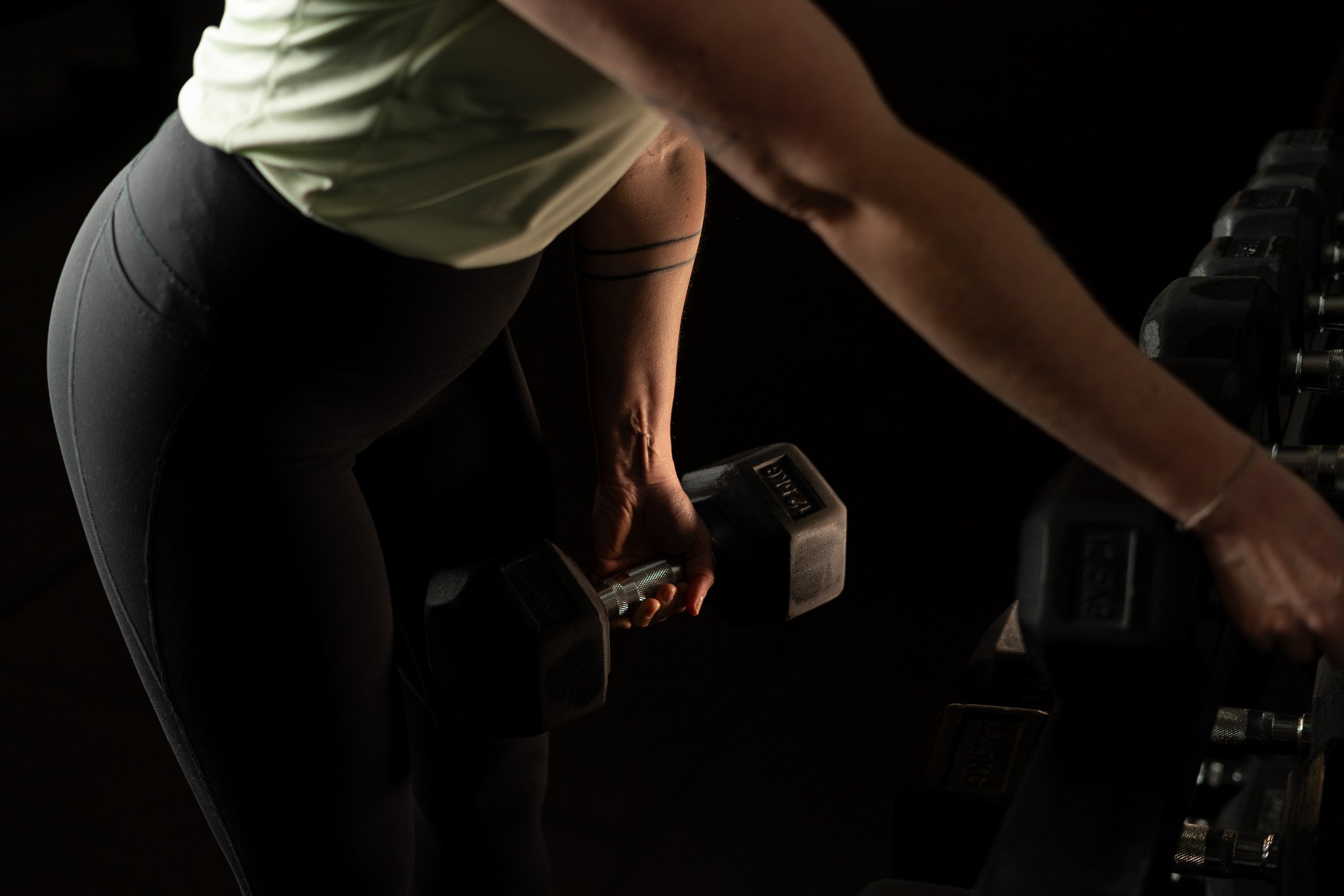 A person in workout clothes, holding a dumbbell in a dark gym environment, with a row of dumbbells visible on a rack.