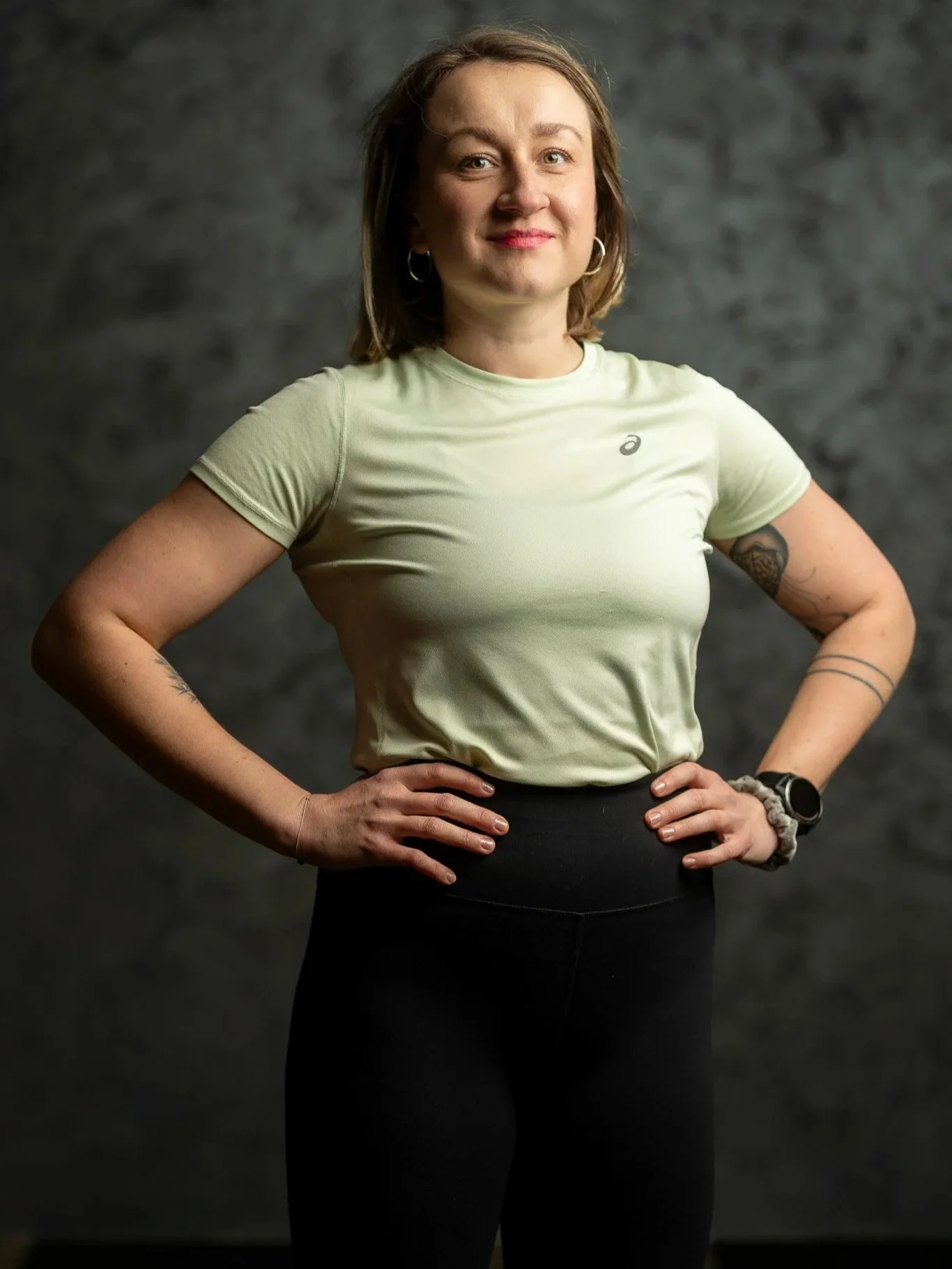A woman with shoulder-length brown hair, wearing a light green athletic shirt and black pants, standing with hands on hips against a dark gray background.