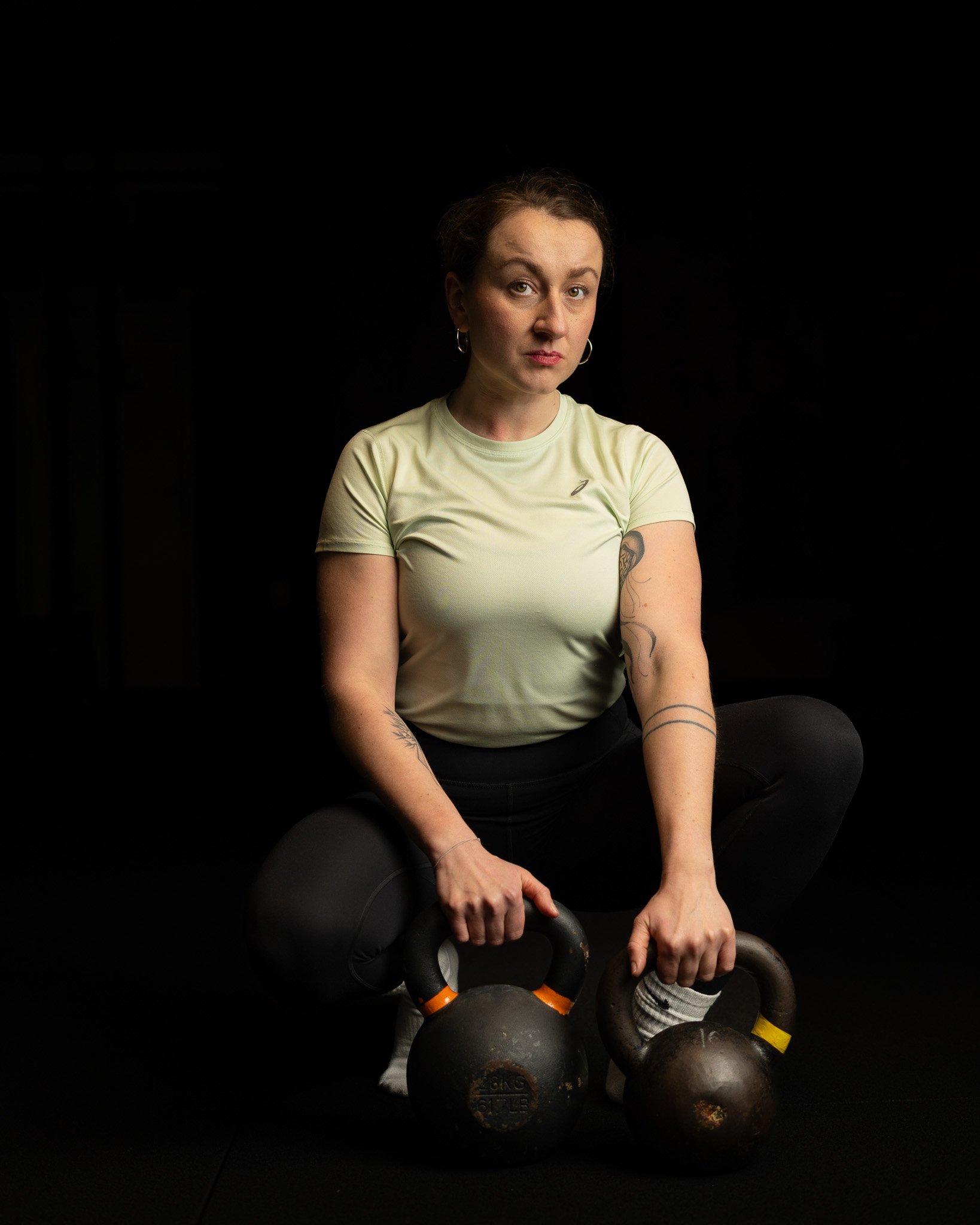 A woman sitting on a black floor surrounded by kettlebells, holding one kettlebell in each hand, against a black background.