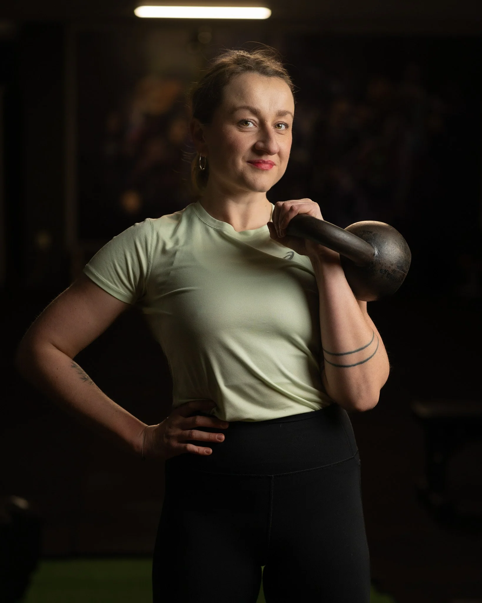 A woman in a light-colored sports shirt holding a kettlebell over her shoulder in a gym or fitness setting.