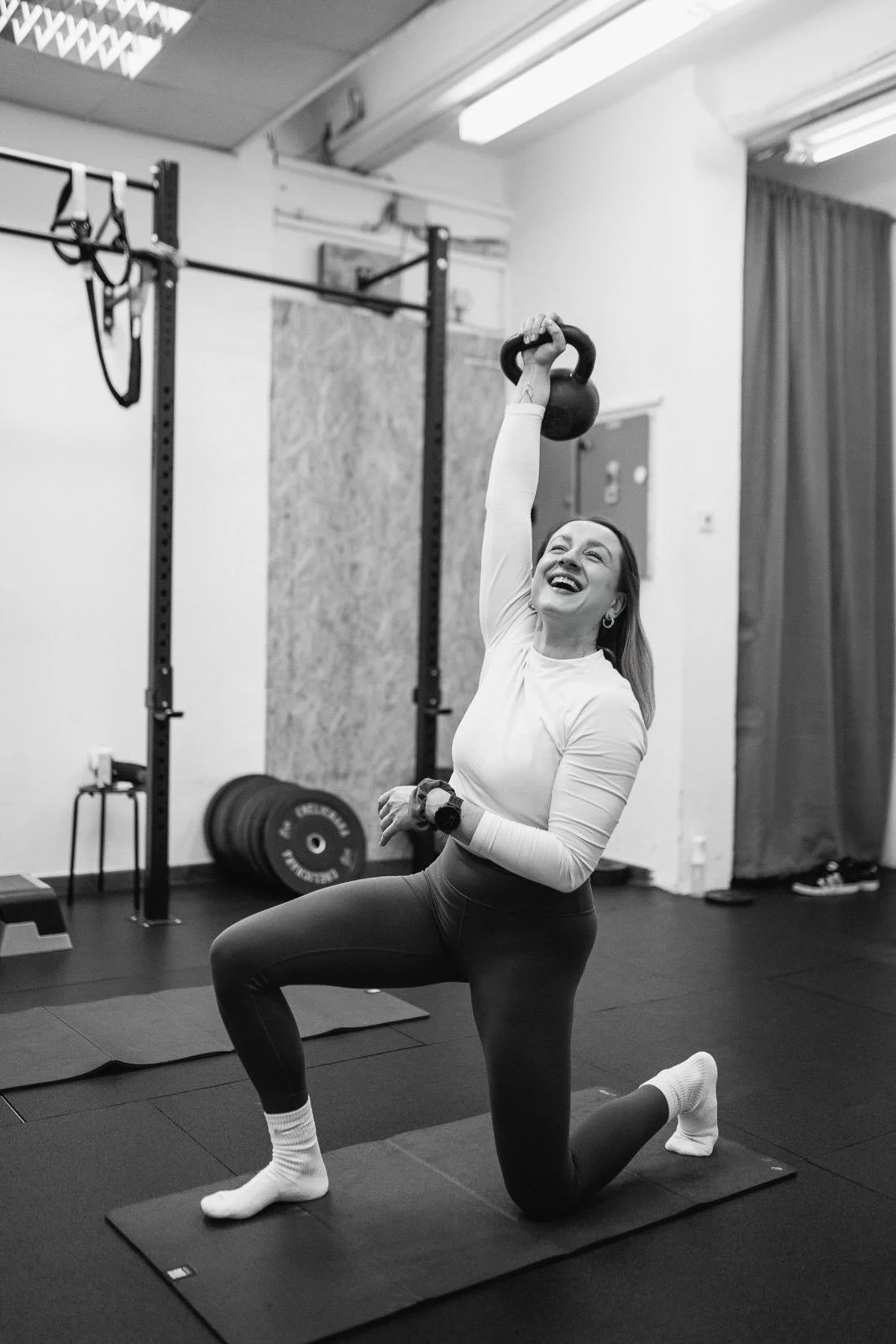 A woman in workout clothes doing a kneeling kettlebell exercise in a gym. She is smiling, holding a kettlebell overhead with one hand, and wearing white sneakers and a long-sleeve shirt.