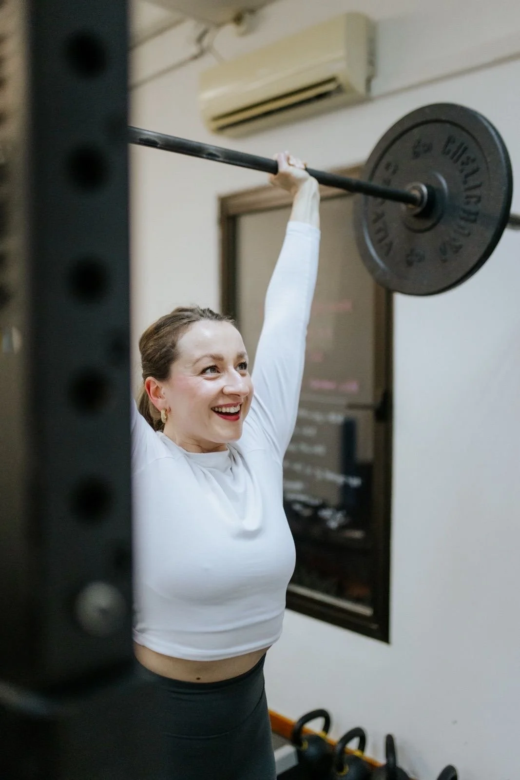 A woman lifting a barbell with a large weight plate in a gym, smiling and looking up.