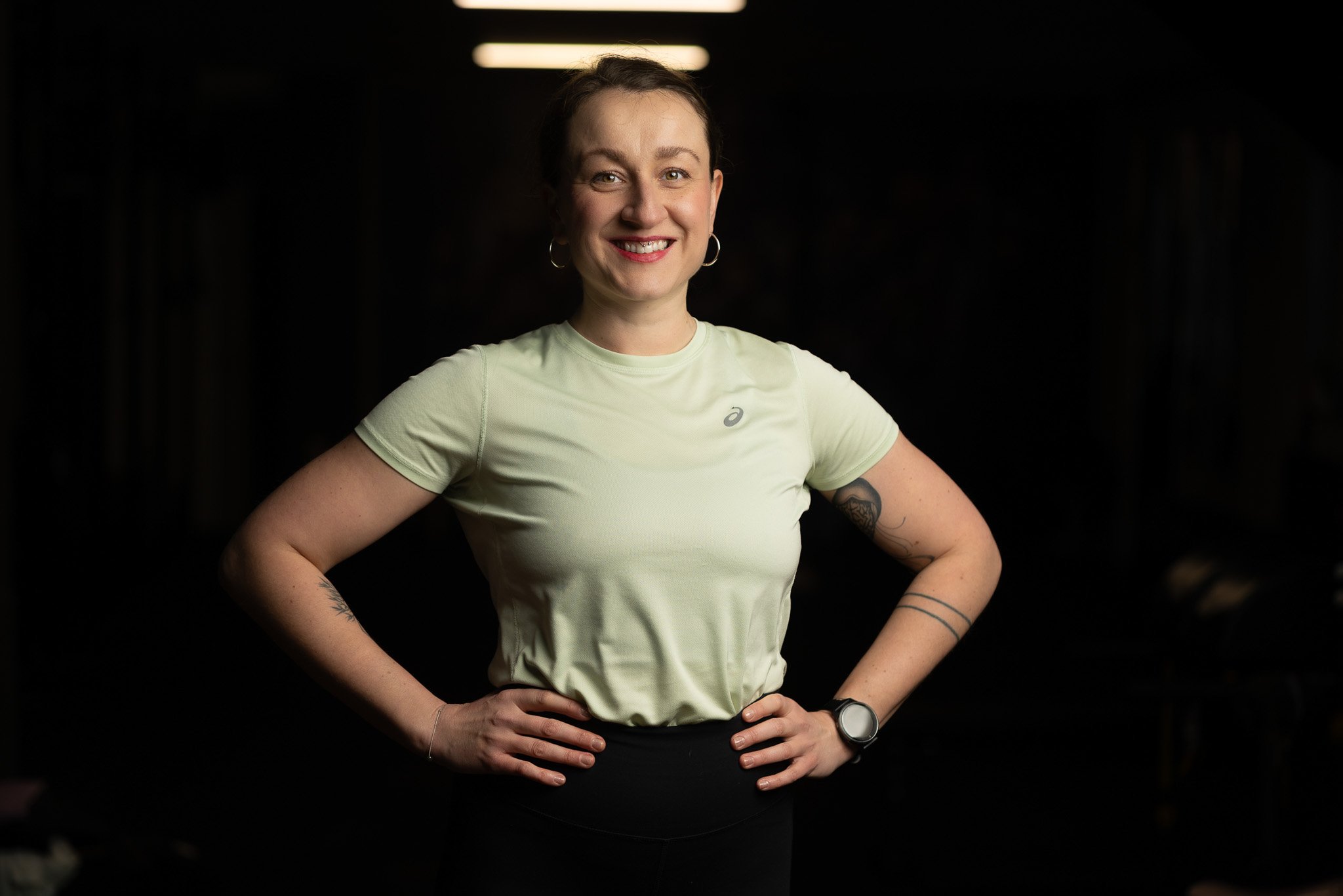 A woman with short hair, earrings, and a watch smiling with hands on hips, wearing a light-colored athletic shirt against a dark background.