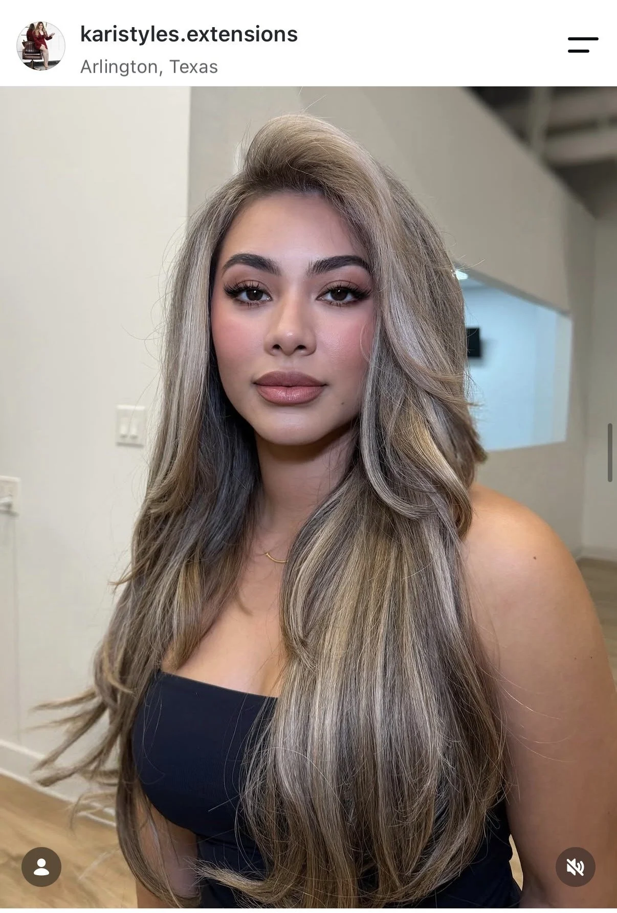 A woman with long, wavy ash blonde hair, wearing makeup, a black strapless top, and a delicate gold necklace, standing indoors in Arlington, Texas.