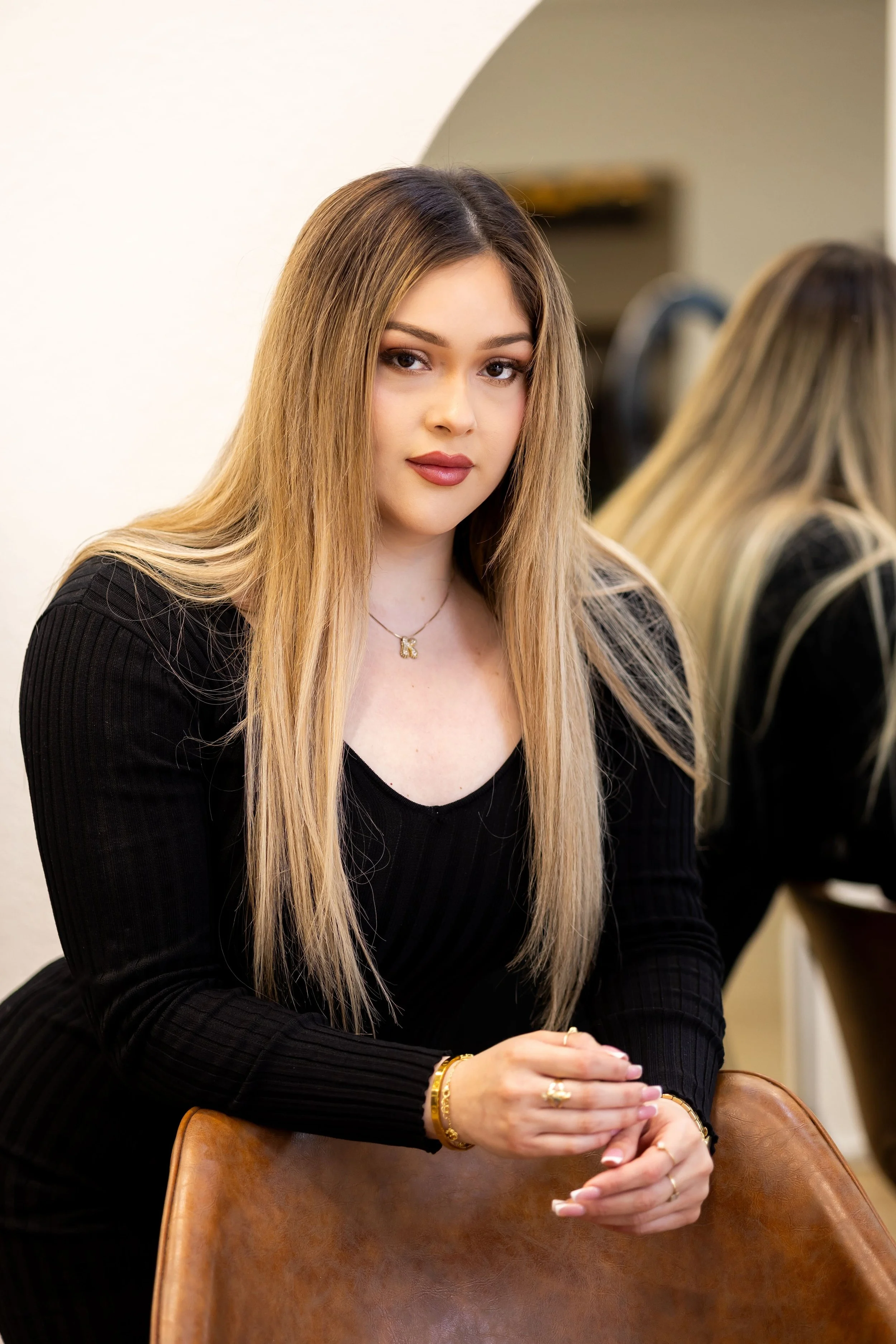 A young woman with long, straight blonde hair, wearing a black ribbed long-sleeve shirt, jewelry including a necklace, bracelets, and rings, sitting on a brown leather chair with her hands clasped, looking at the camera with a neutral expression, in an indoor setting.