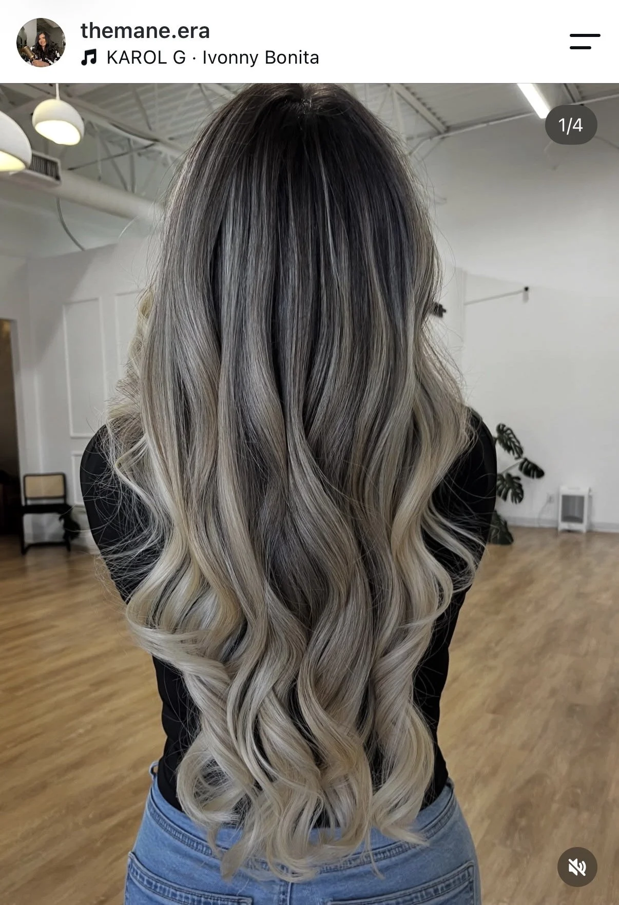 Back view of a woman with long wavy hair in ombre shades of black, gray, and blonde, wearing a black top and blue jeans, in a hair salon with wooden floors and white walls.
