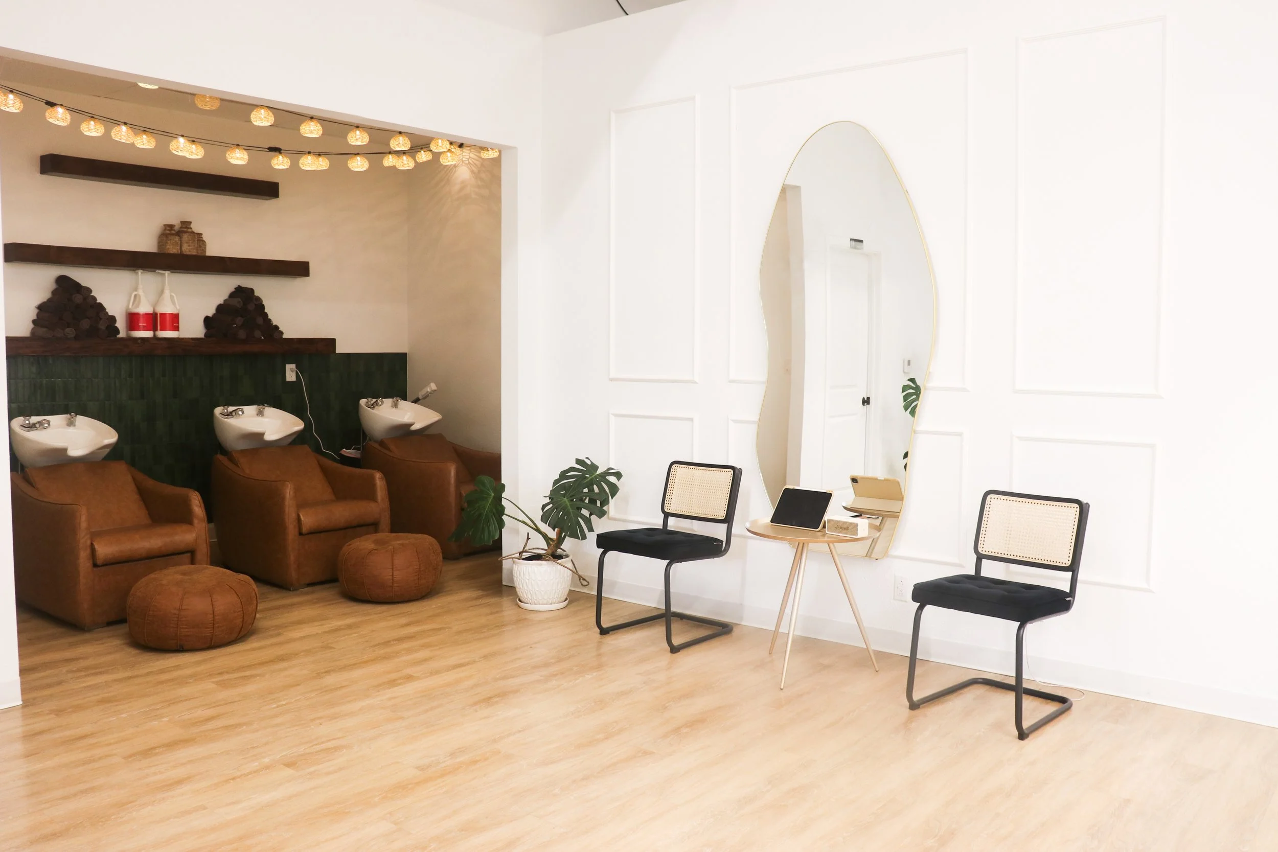 A waiting area with brown leather chairs and poufs, black and beige chairs, a round wooden table with electronic devices, potted plants, and a tall mirror with a gold frame. The background features a white wall with decorative paneling.