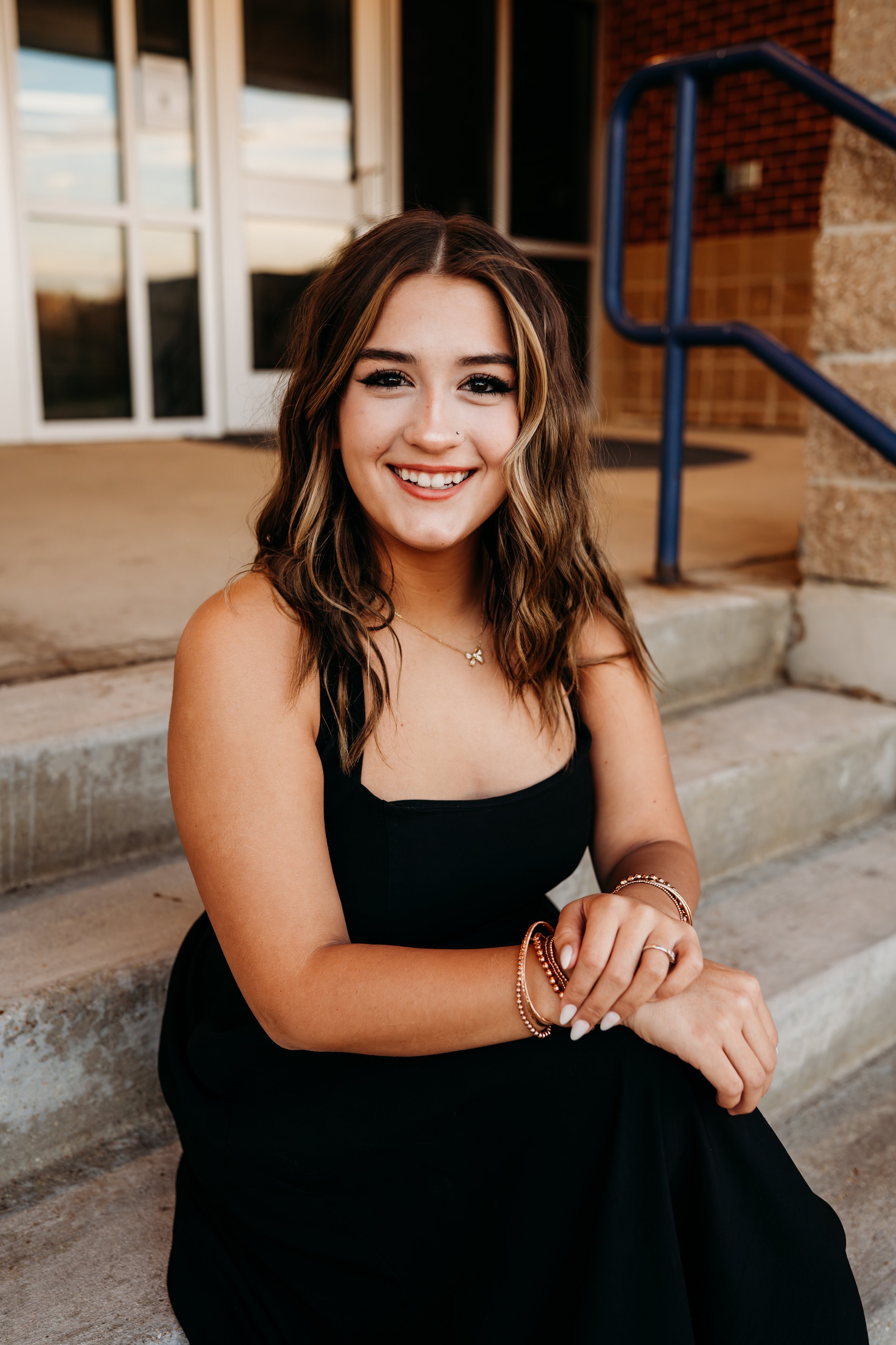 A young woman with wavy brown hair, wearing a black dress and jewelry, smiling and sitting on concrete steps outside a building with glass doors and brick walls.