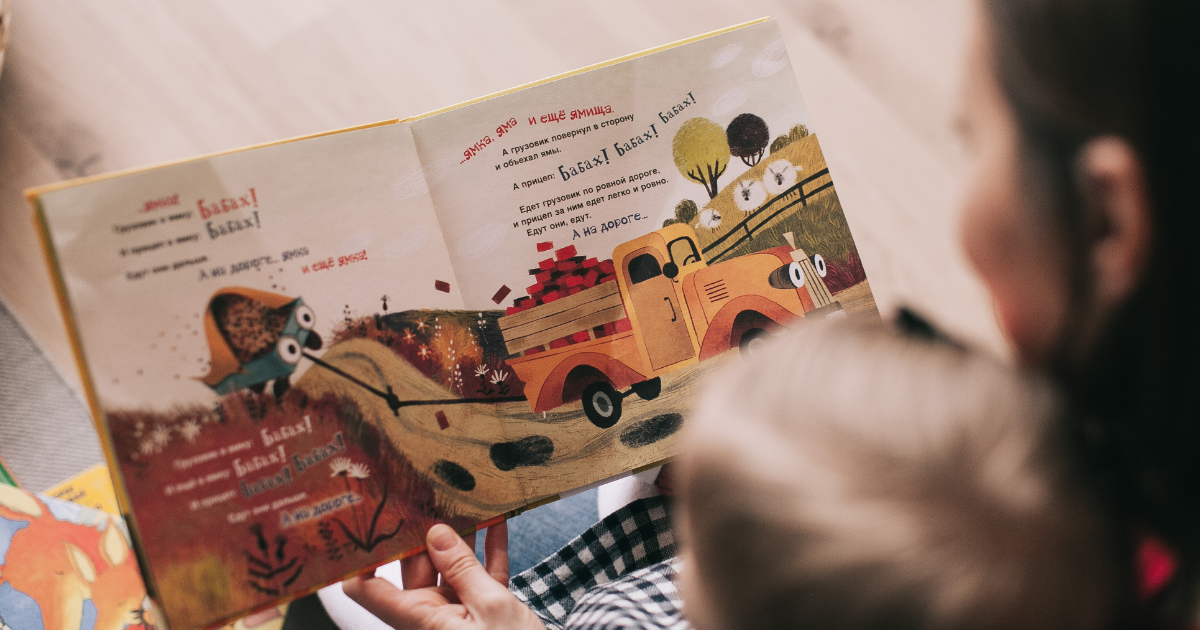A child and an adult reading a colorful children's book featuring a cartoon tractor pulling a wagon of bricks and animals in a countryside setting.