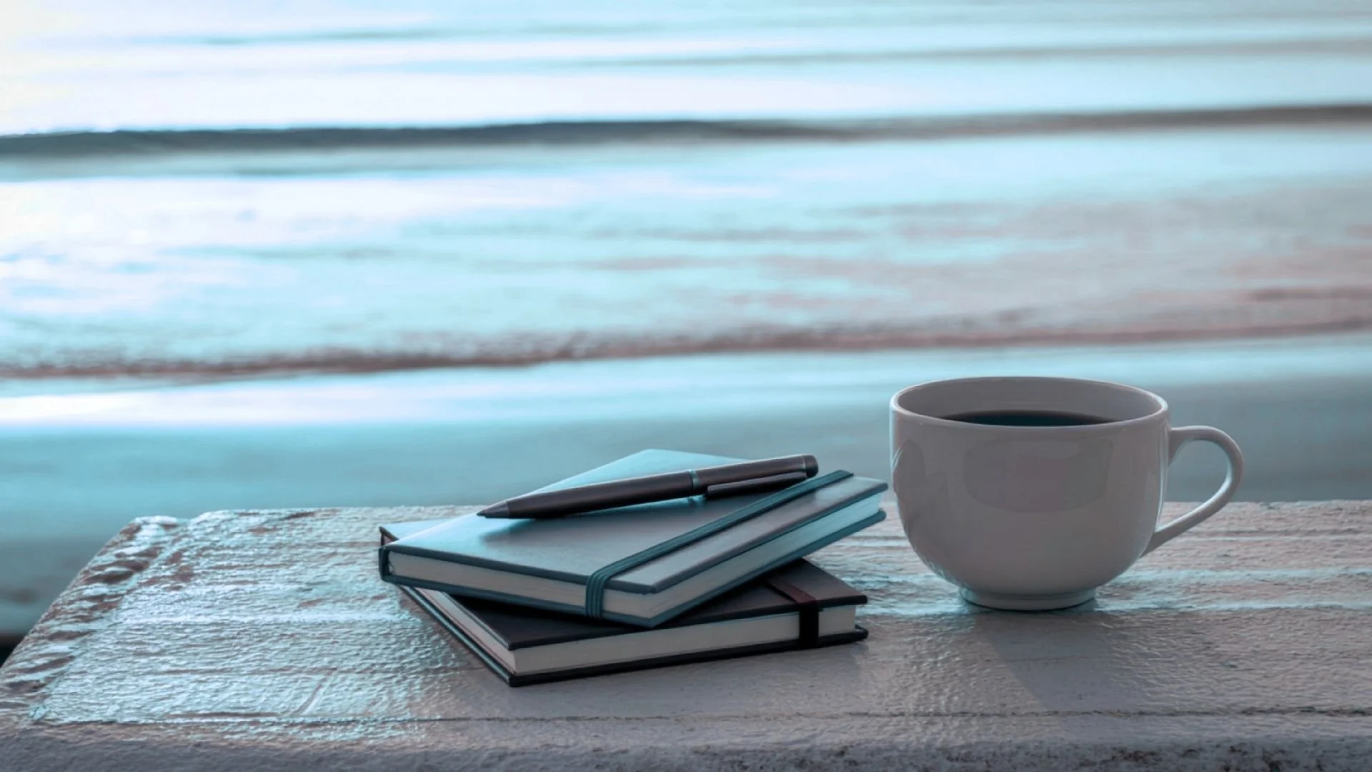 A cup of black coffee, stacked notebooks with pens, placed on a table near a window overlooking a beach with ocean waves.