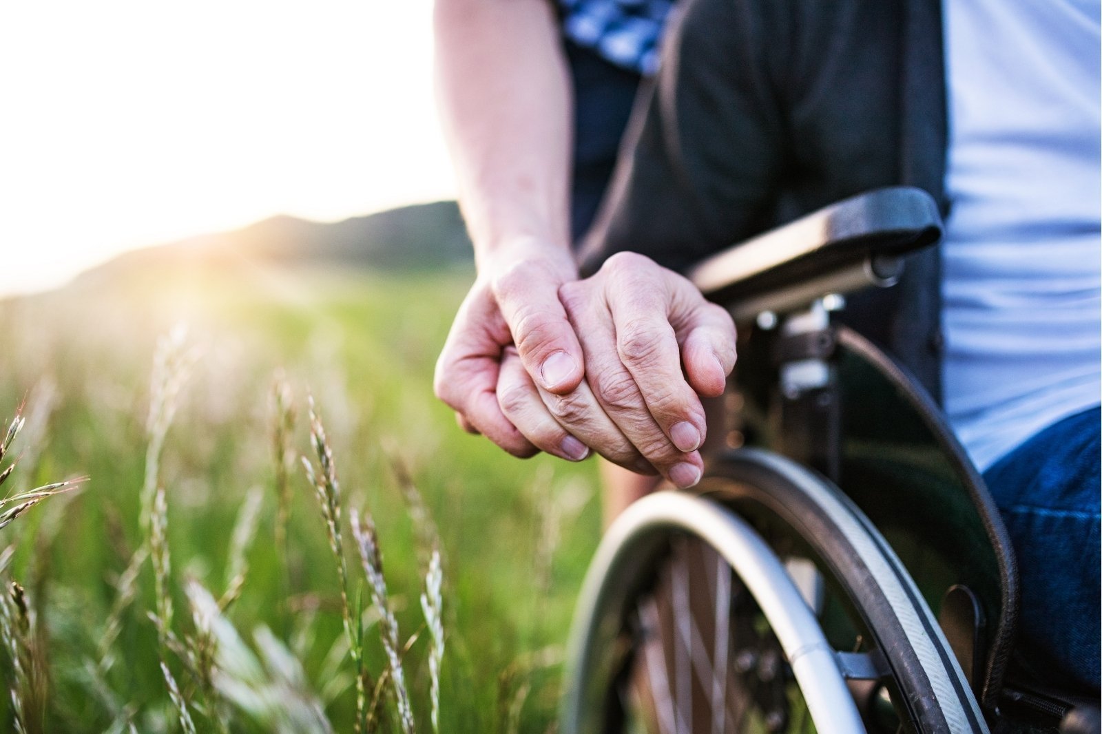 A person gently holds the hand of Mike Shultz while he sits in his wheelchair outdoors, symbolizing unwavering love, support, and hope.