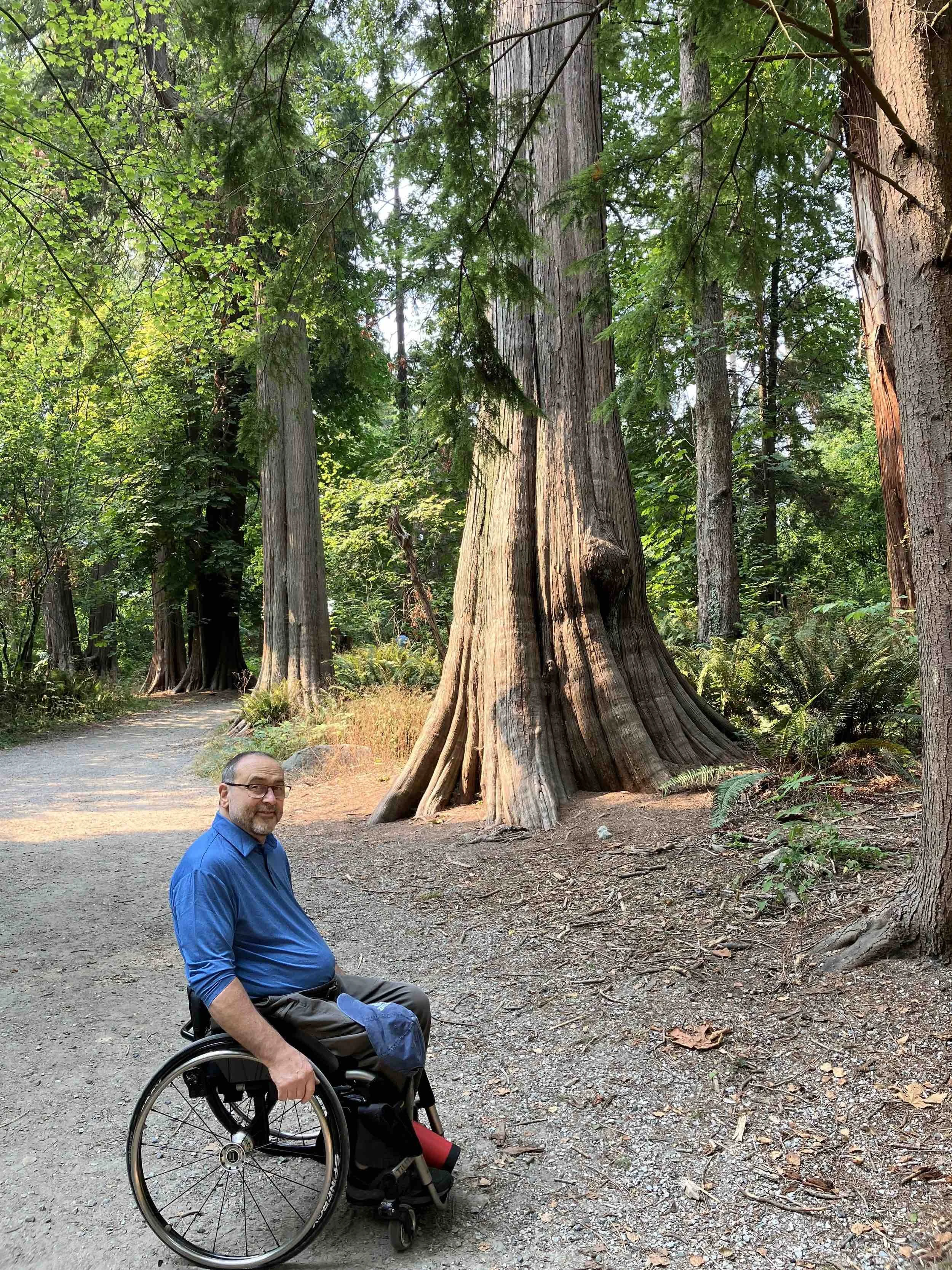 Mike Shultz, author of You Can't Stop Love, smiles while seated in his wheelchair beside towering trees during an outdoor adventure.