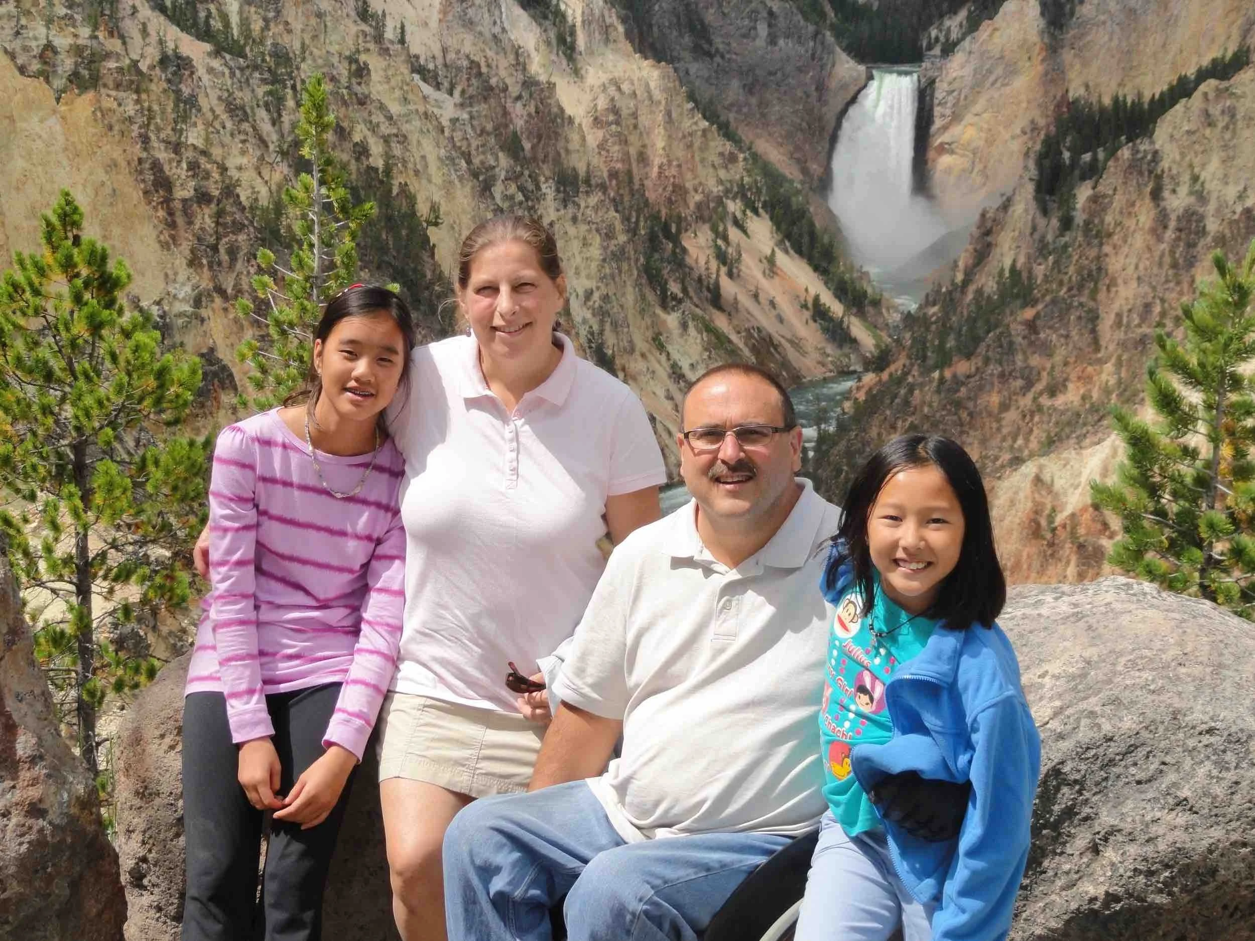 Mike Shultz poses with his family at Yellowstone National Park, with a waterfall and canyon in the background.