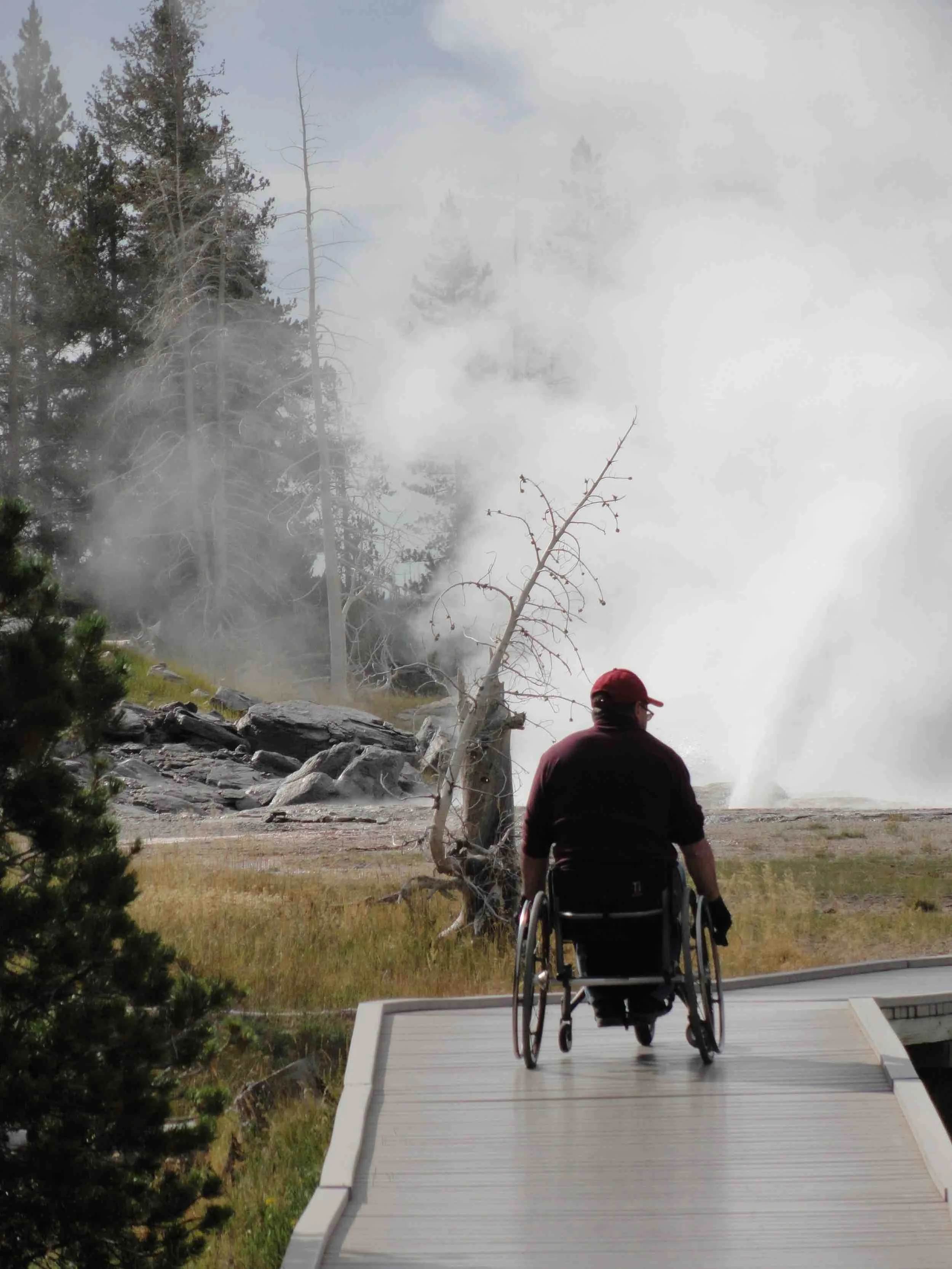Mike Shultz explores a Yellowstone National Park boardwalk in his wheelchair near a steaming geothermal feature.