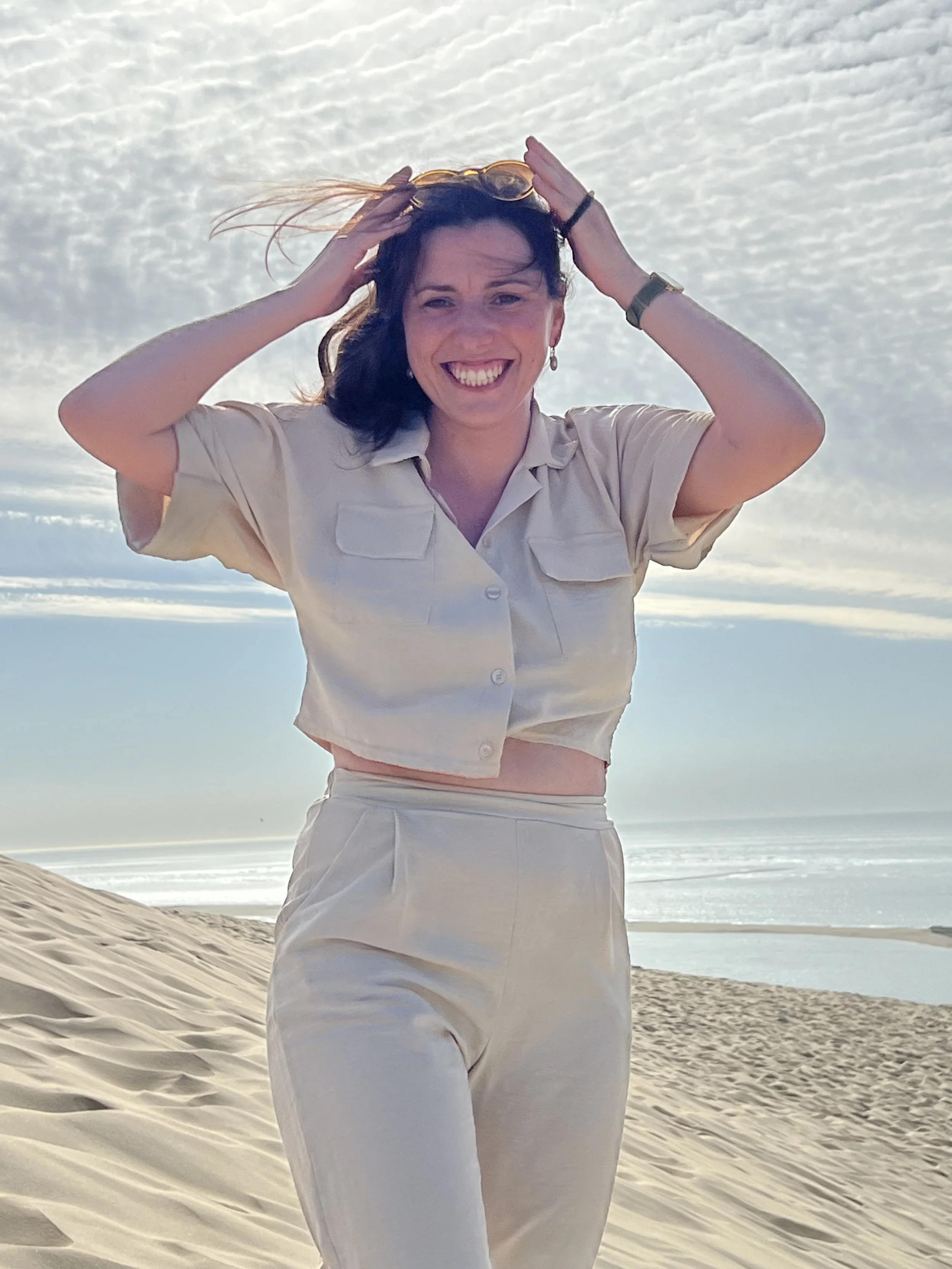 Une femme souriante debout sur la plage, portant des vêtements blancs, avec le ciel et la mer en arrière-plan.