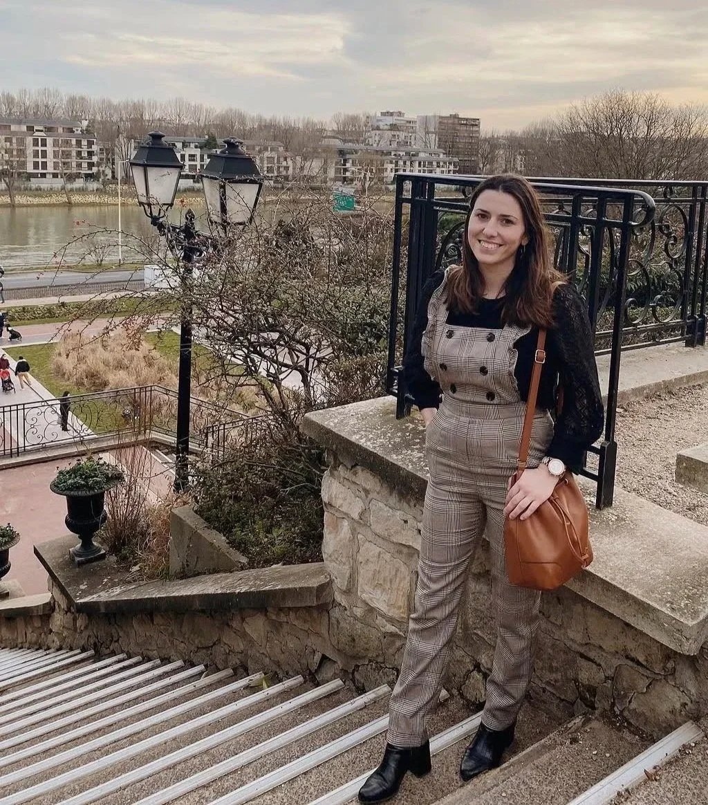 Femme souriante tenant un sac en cuir marron, posant sur un escalier en pierre près d'une rivière, avec des bâtiments modernes en arrière-plan, en fin d'après-midi ou début de soirée.