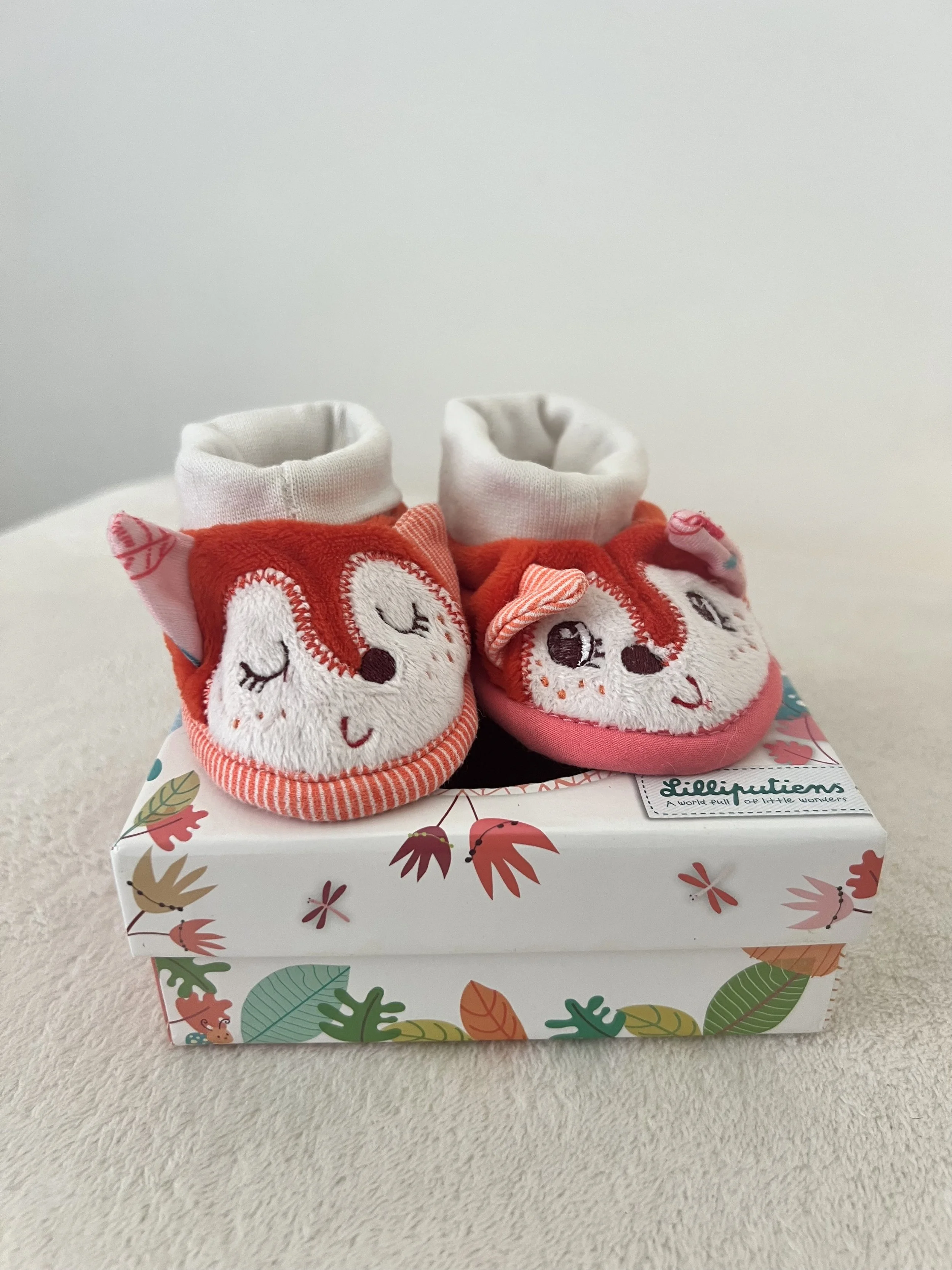 Paquet de chaussons pour bébé avec des visages d'animaux souriants, posés sur une boîte décorée de motifs de feuilles colorées.