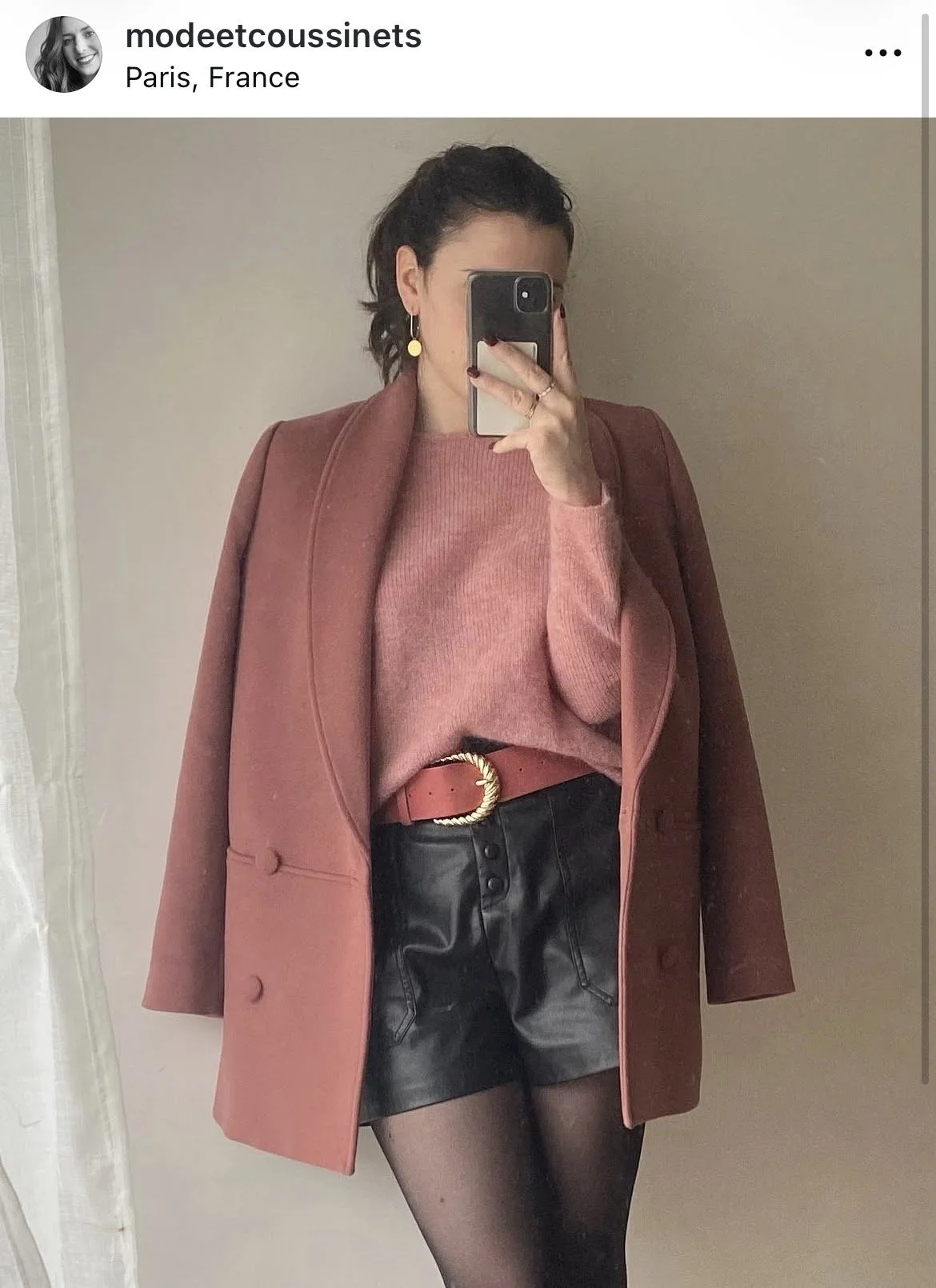 Une femme habillée d'un manteau rose Sézane, un pull rose Sézane, un short en cuir noir Only, porte des collants noirs Calzedonia, et un ceinture rose Grace and Mila avec une boucle dorée, prend une photo selfie dans un miroir.