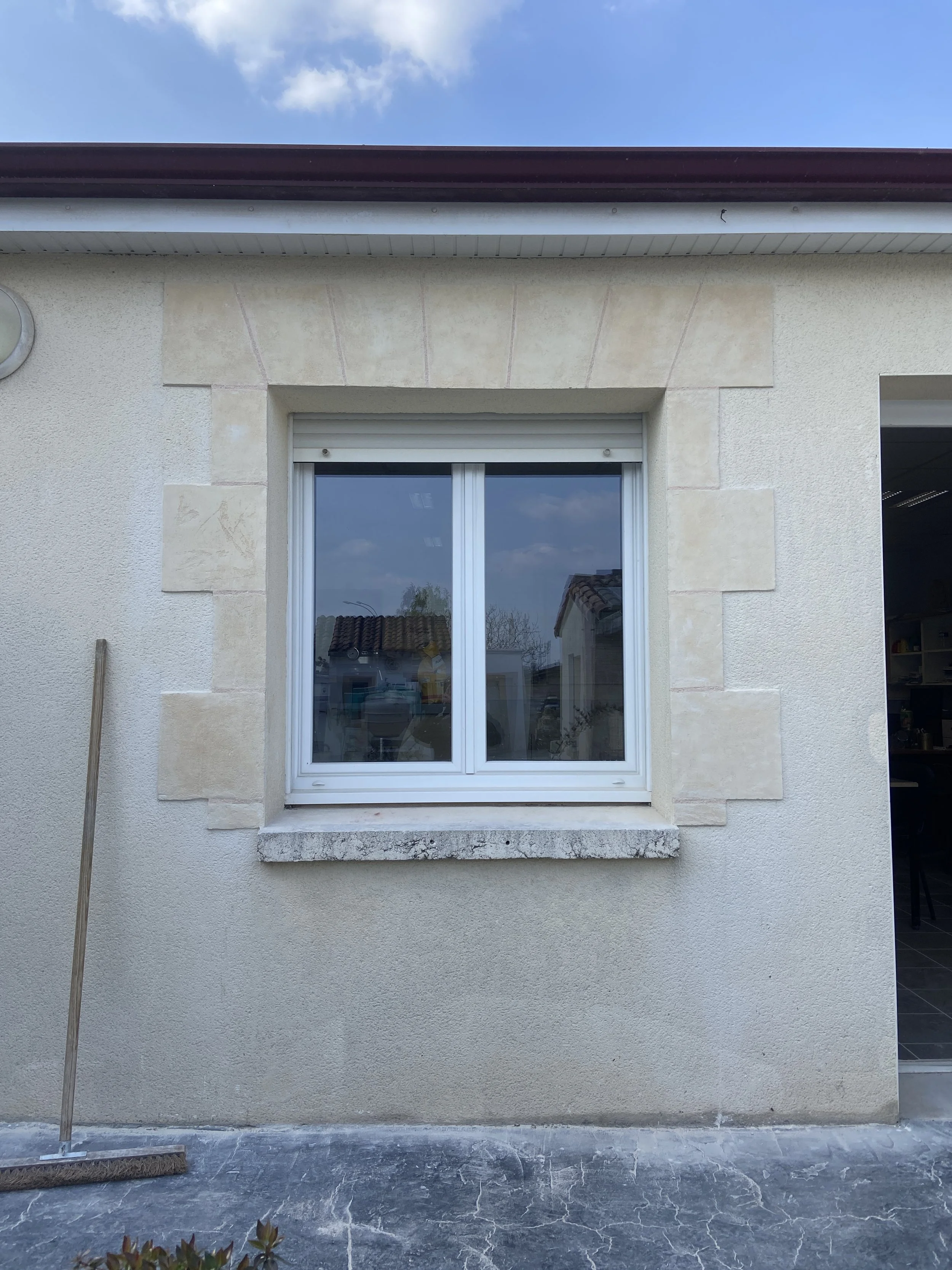 Fenêtre blanche encadrée de pierre sur un mur beige, avec un ciel bleu et quelques nuages, et un sol en pierre.