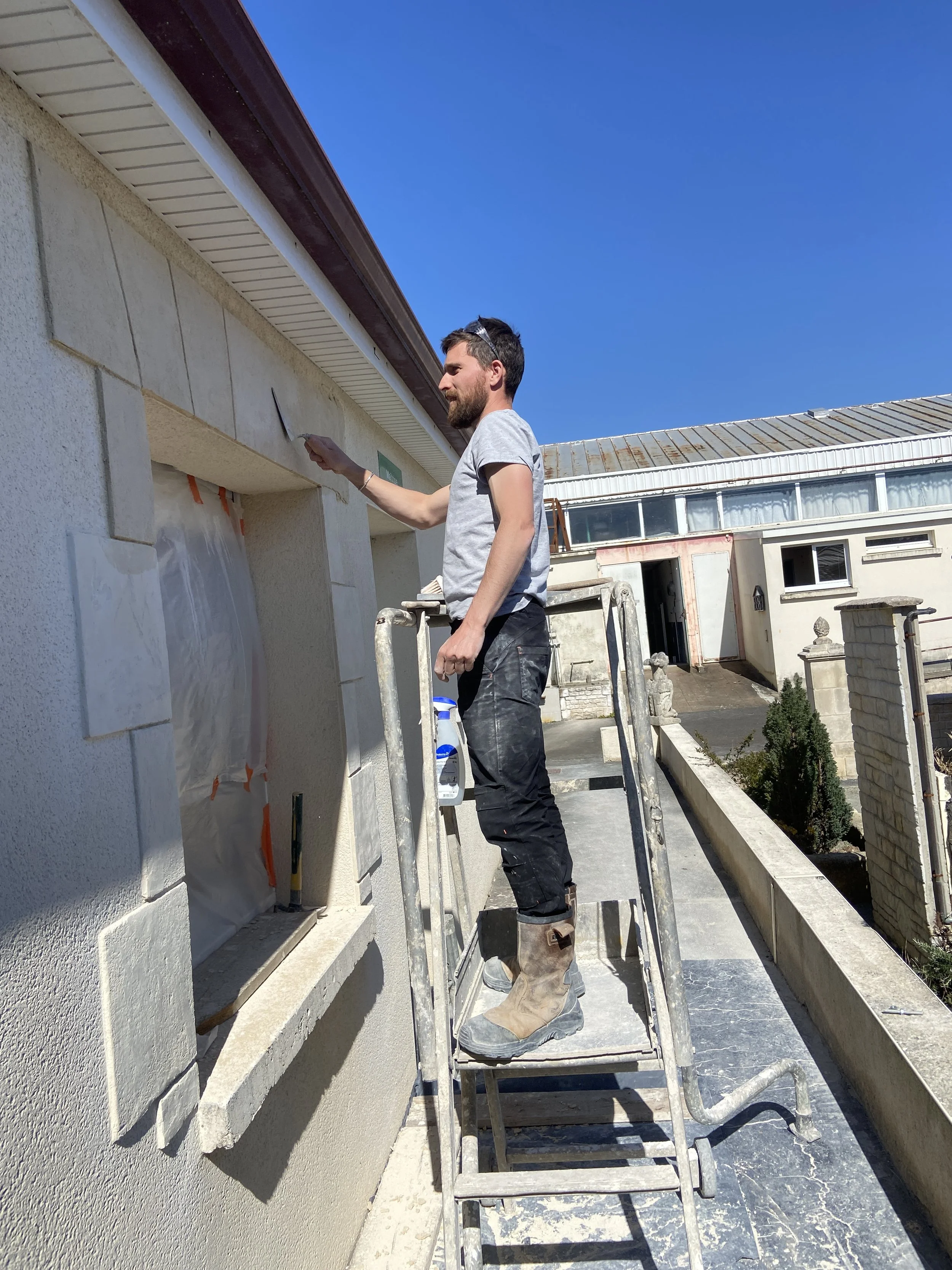 Un homme avec une barbe, portant un t-shirt gris et des bottes de chantier, se tient sur une échelle et peint la façade d'un bâtiment en blanc sous un ciel bleu clair.