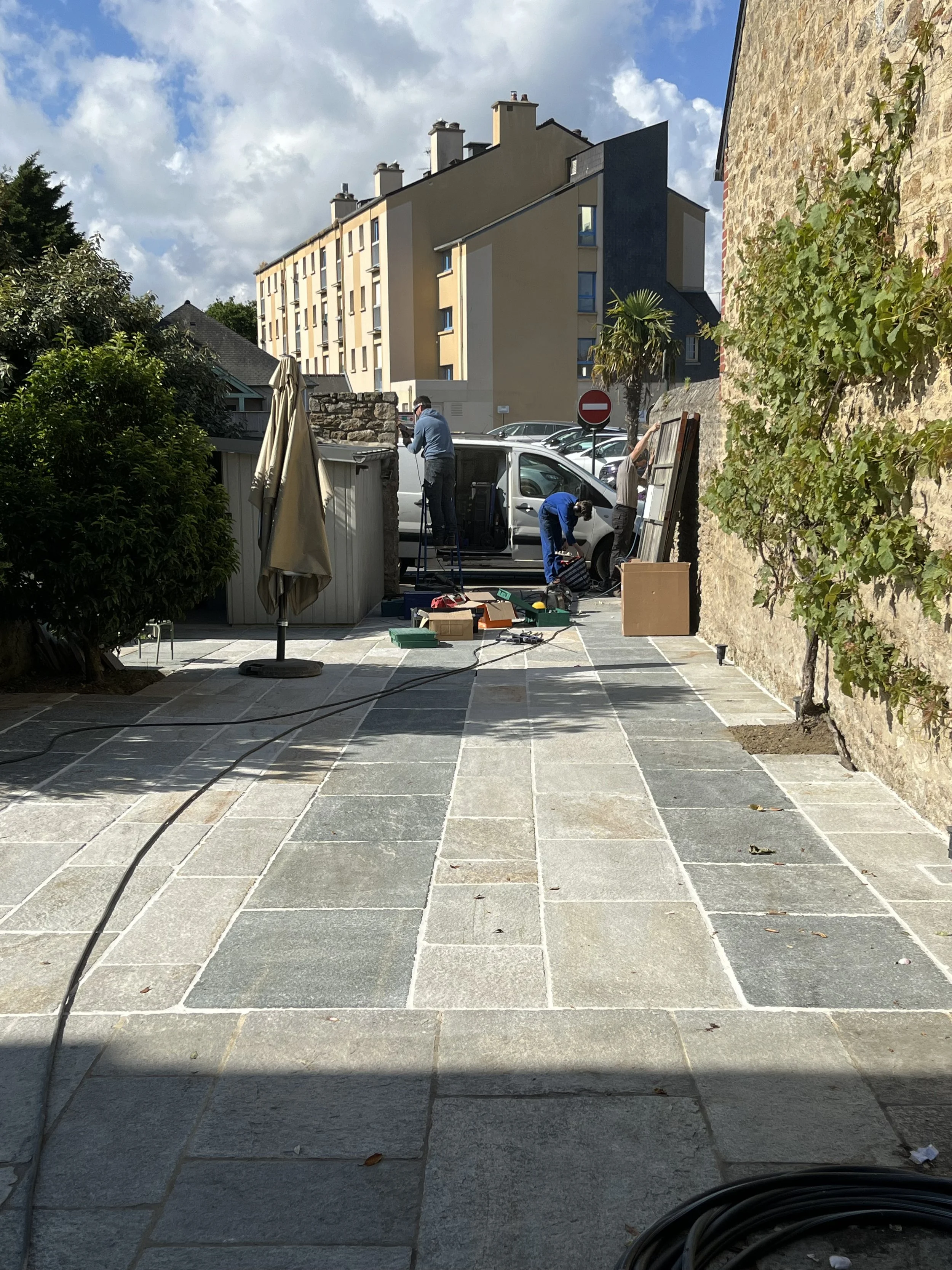 Des ouvriers installent ou réparent un carrelage extérieur dans une cour pavée, avec un véhicule blanc garé et un mur en pierre sur le côté droit.
