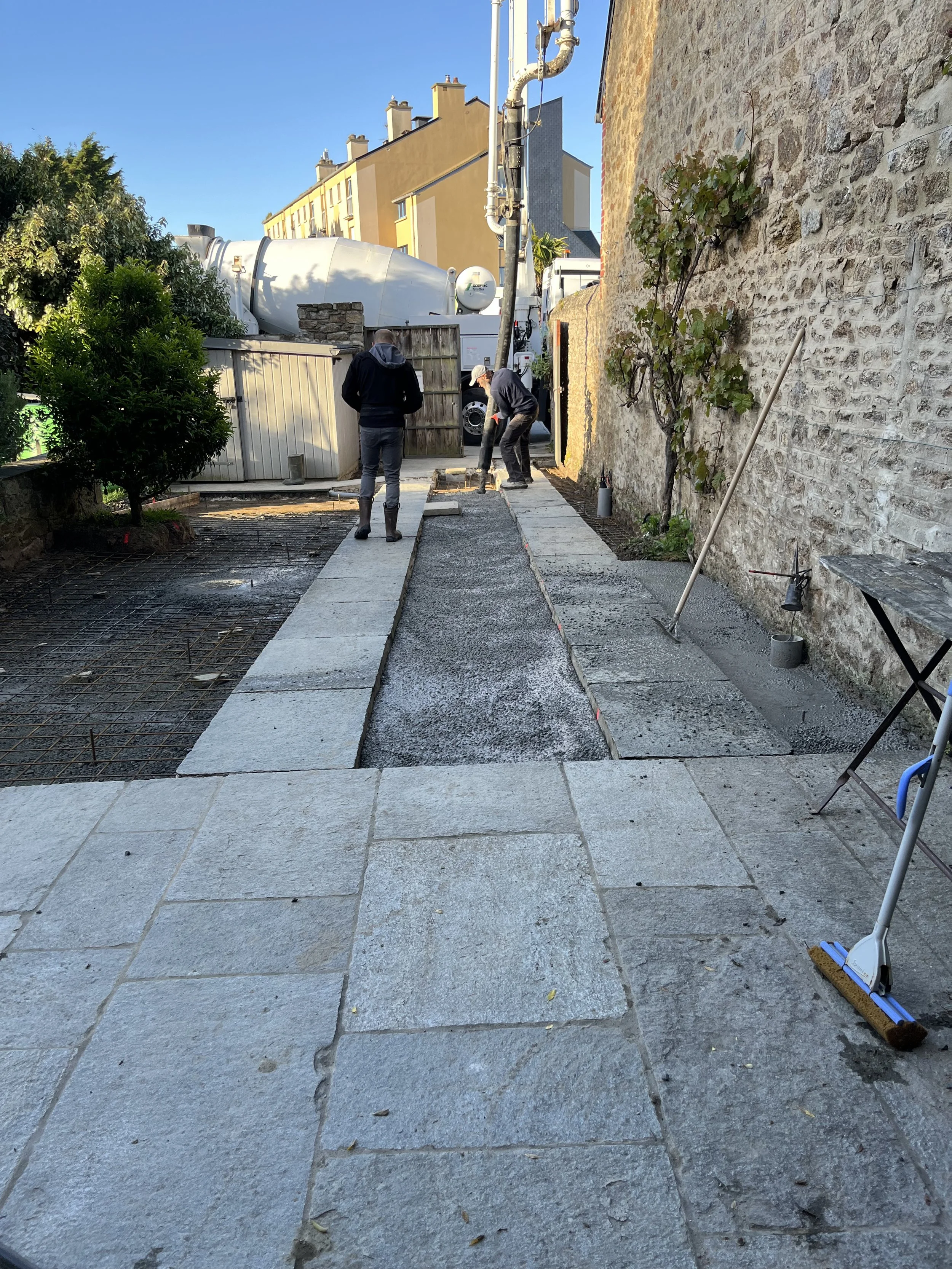 Deux personnes qui travaillent à la construction ou à la rénovation d'une terrasse en pavés gris clair, avec des outils de nettoyage et des matériaux de construction posés au sol, à côté d'un mur en pierre et d'une clôture en bois.