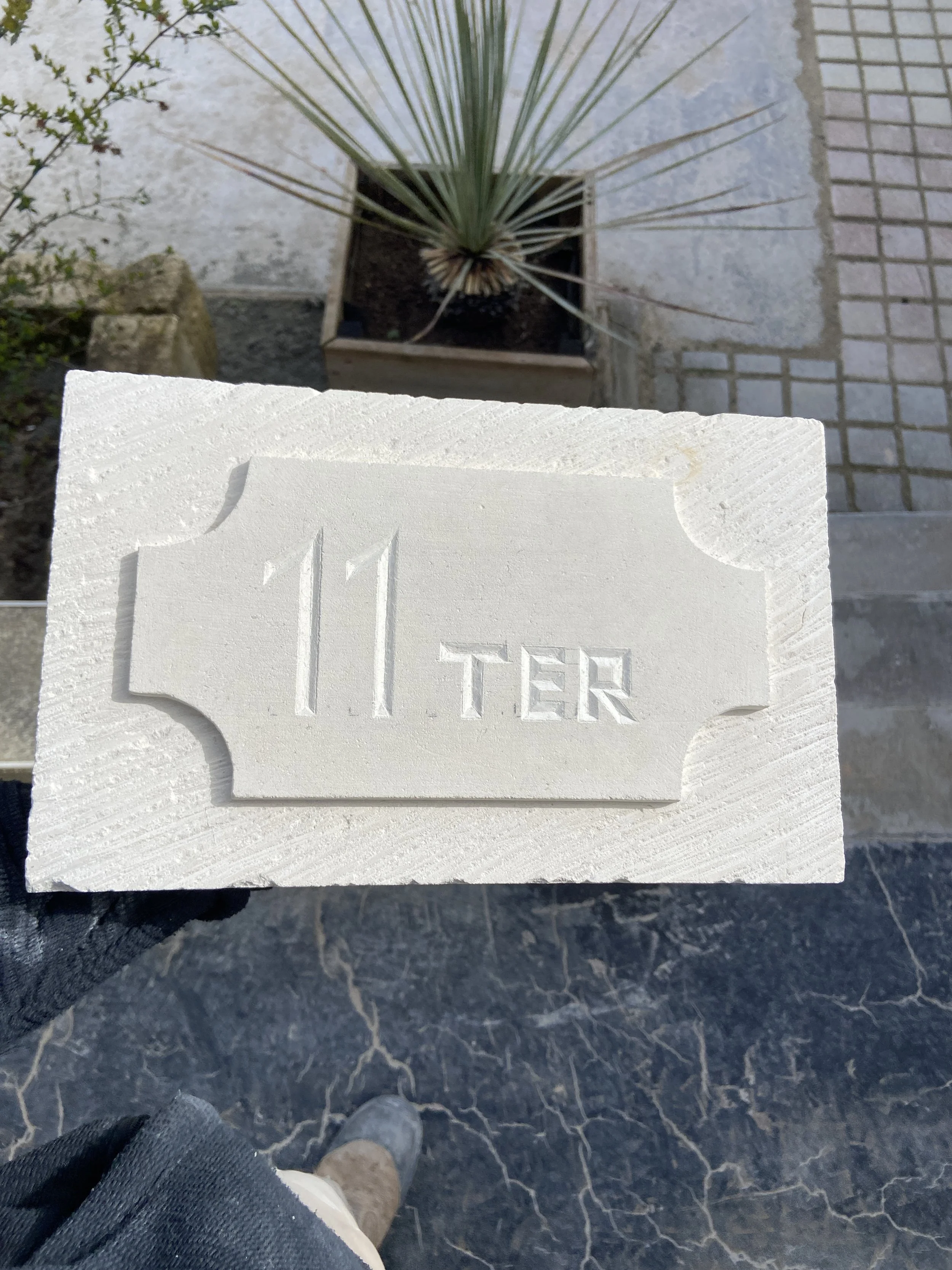 Photo vue du haut d'une personne portant des chaussures beige, des jeans déchirés, regardant en bas un sol noir avec du marbre blanc et une bordure en pavé. En face, un mur avec une plaque blanche gravée avec le texte 'Il TERRA'.