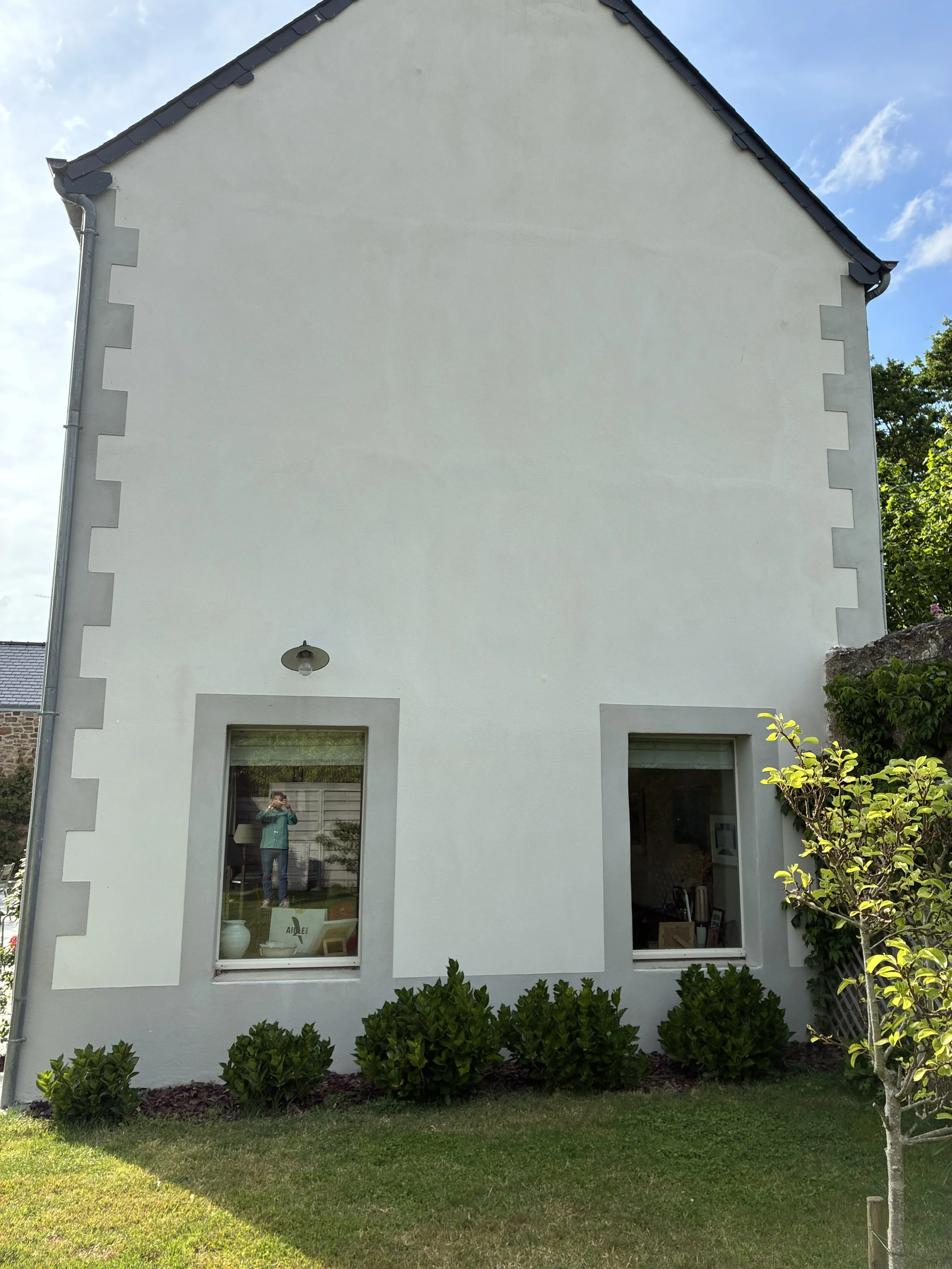 Façade blanche d'une maison vue de face avec deux grandes fenêtres, un jardin avec buissons et un arbre, et un ciel bleu avec quelques nuages.
