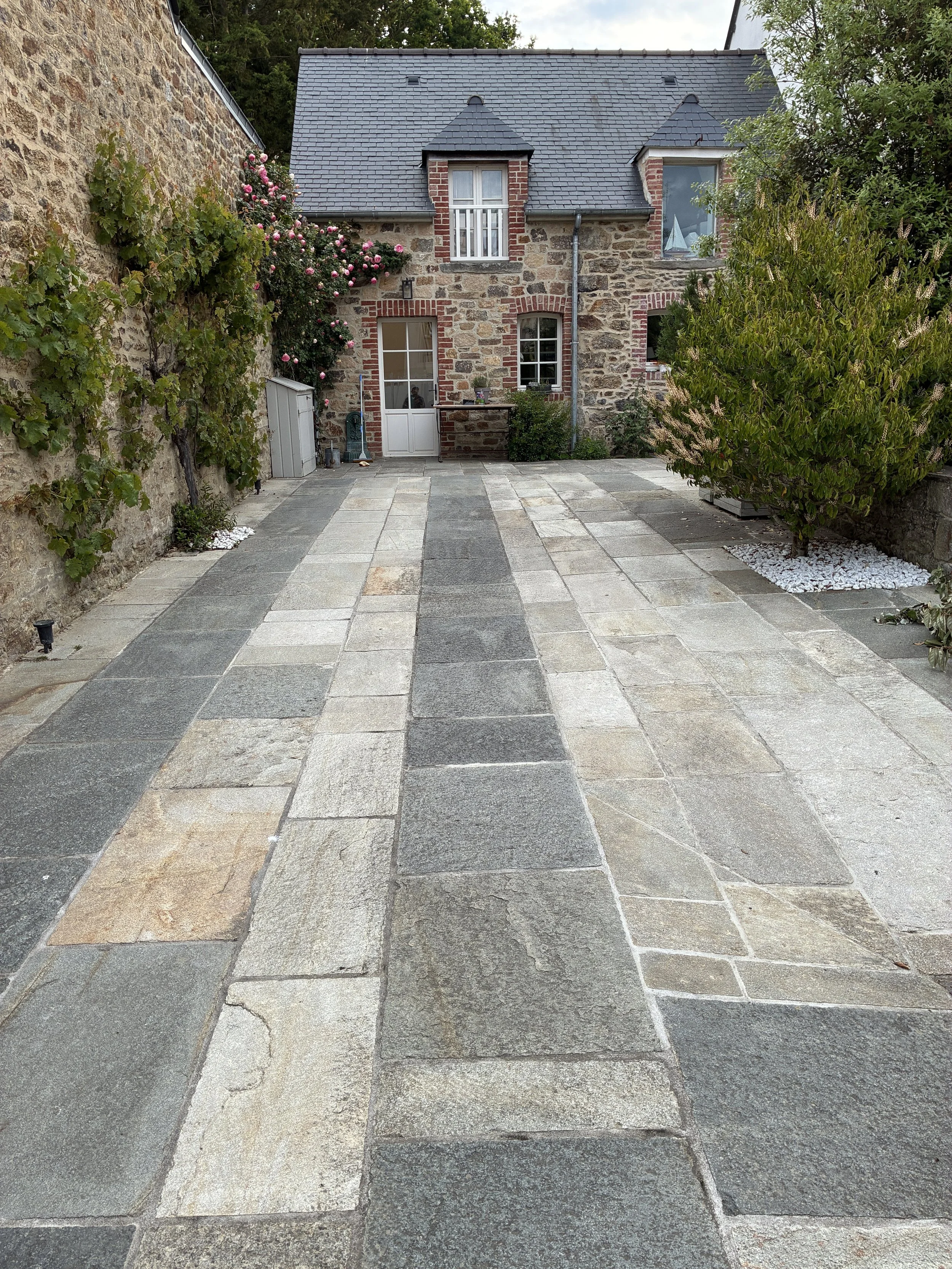 Cour pavée devant une maison en pierre avec toiture en ardoise, entourée de végétation et de fleurs.