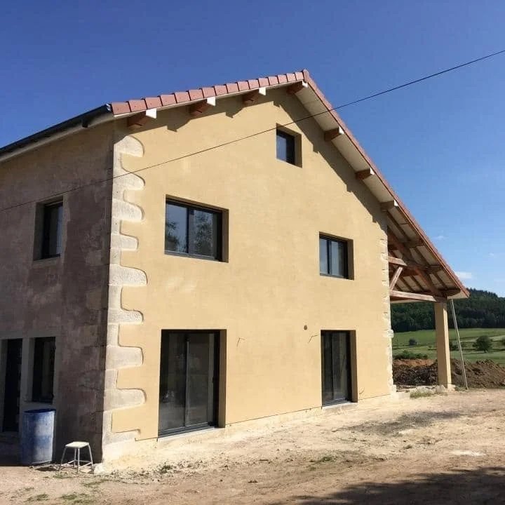 Maison en construction avec murs en plâtre jaune et fenêtres modernes, sous un toit en tuiles rouges, dans un environnement rural
