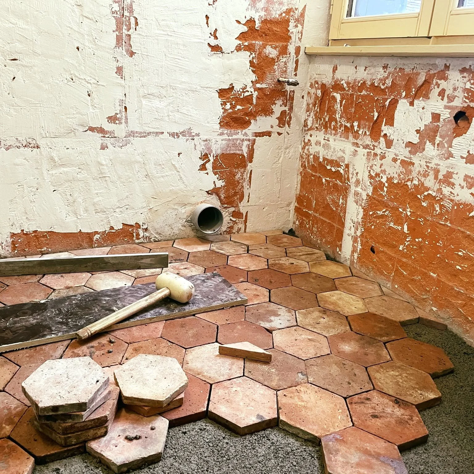 Une pièce en rénovation avec un mur en brique et une moquette en cours de pose. Sur le sol, des carreaux en forme d'hexagone, certains empilés, et un marteau repose sur une planche