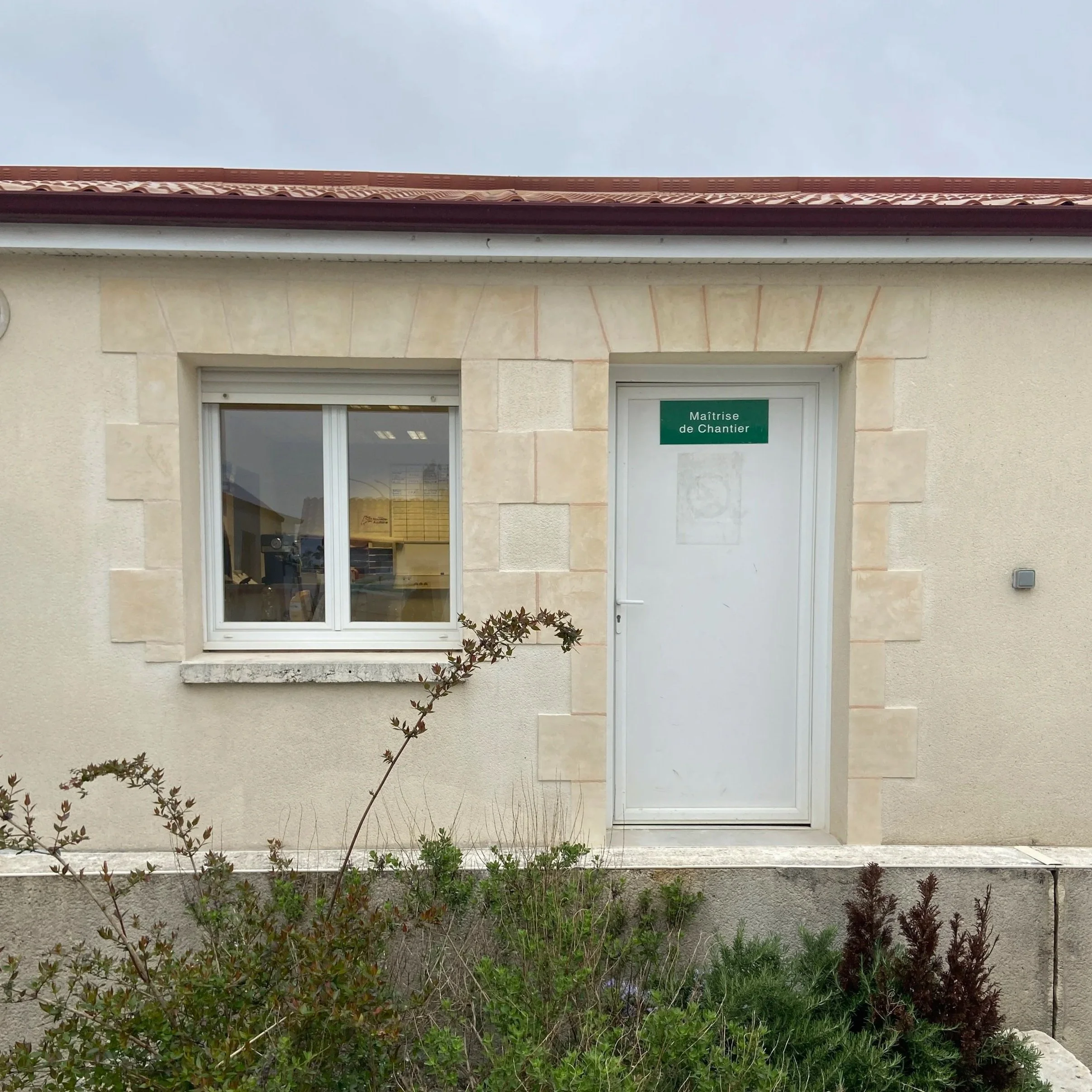 Vue de la façade d'un bâtiment résidentiel avec une porte et une fenêtre, entouré de plantes et de petits arbustes, avec un ciel clair en arrière-plan.