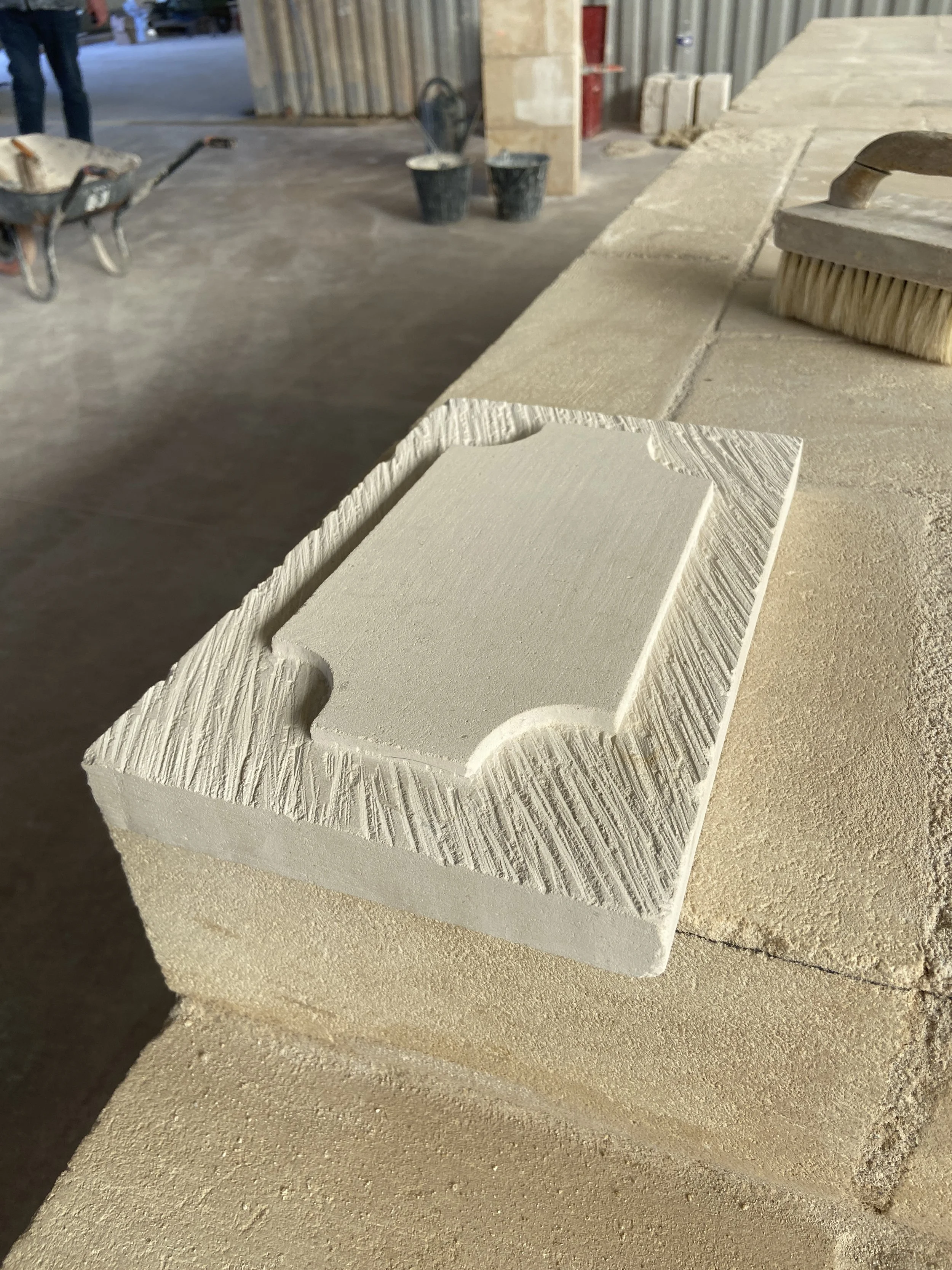 Bloc de pierre ou de matériau de construction avec forme rectangulaire et une cavité ronde dans le coin supérieur gauche, placé sur une surface de sable ou de ciment, avec un pinceau à droite.