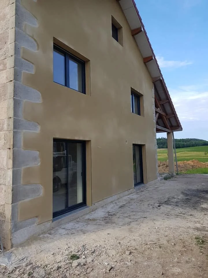 Façade d'une maison avec des fenêtres et un sol en terre, en cours de construction, dans un environnement rural.