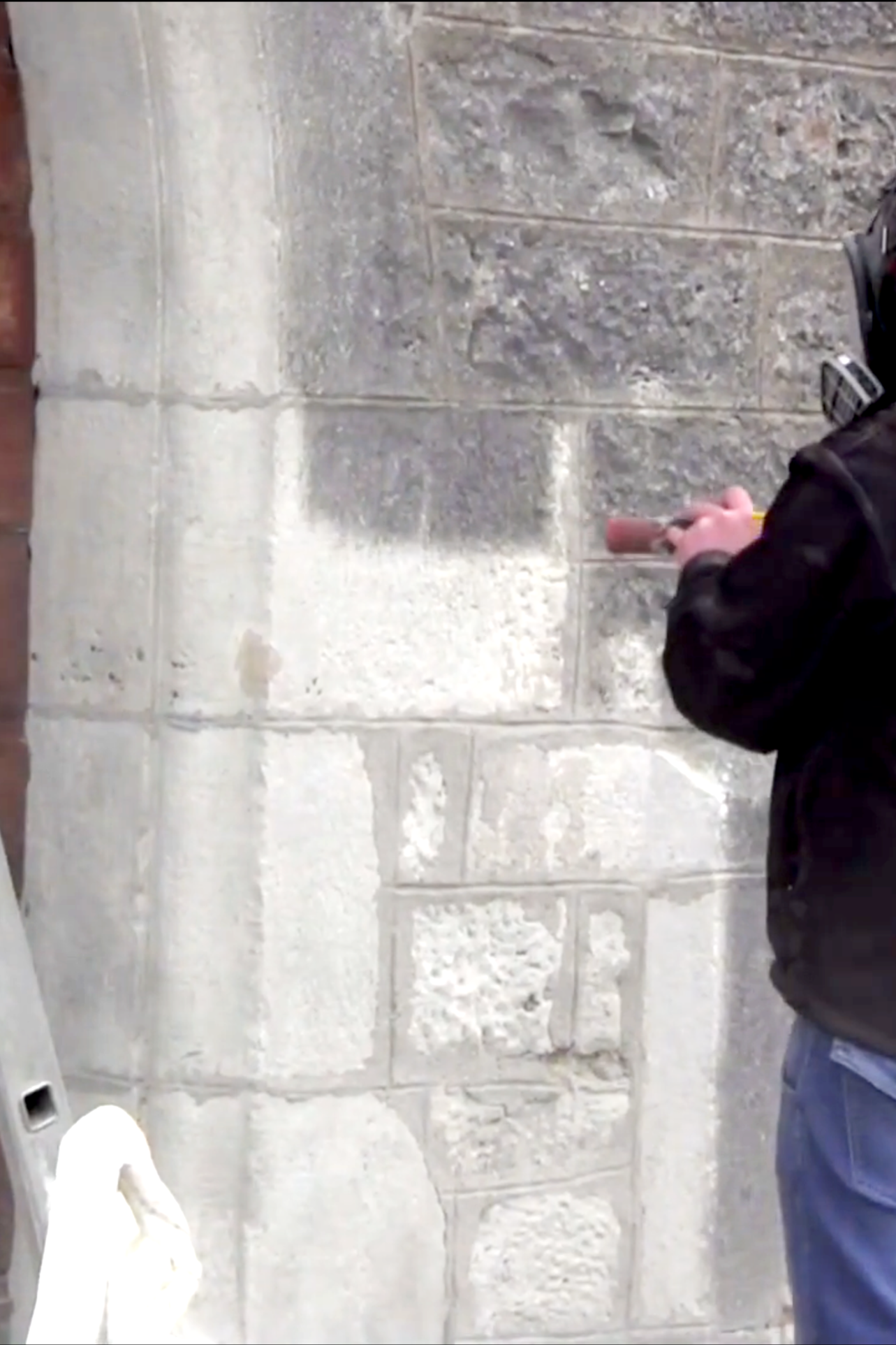 Une personne en veste noire nettoie un mur en pierre avec une brosse. La photo est prise dans une pièce avec un sol en marbre et une toilette blanche en premier plan.