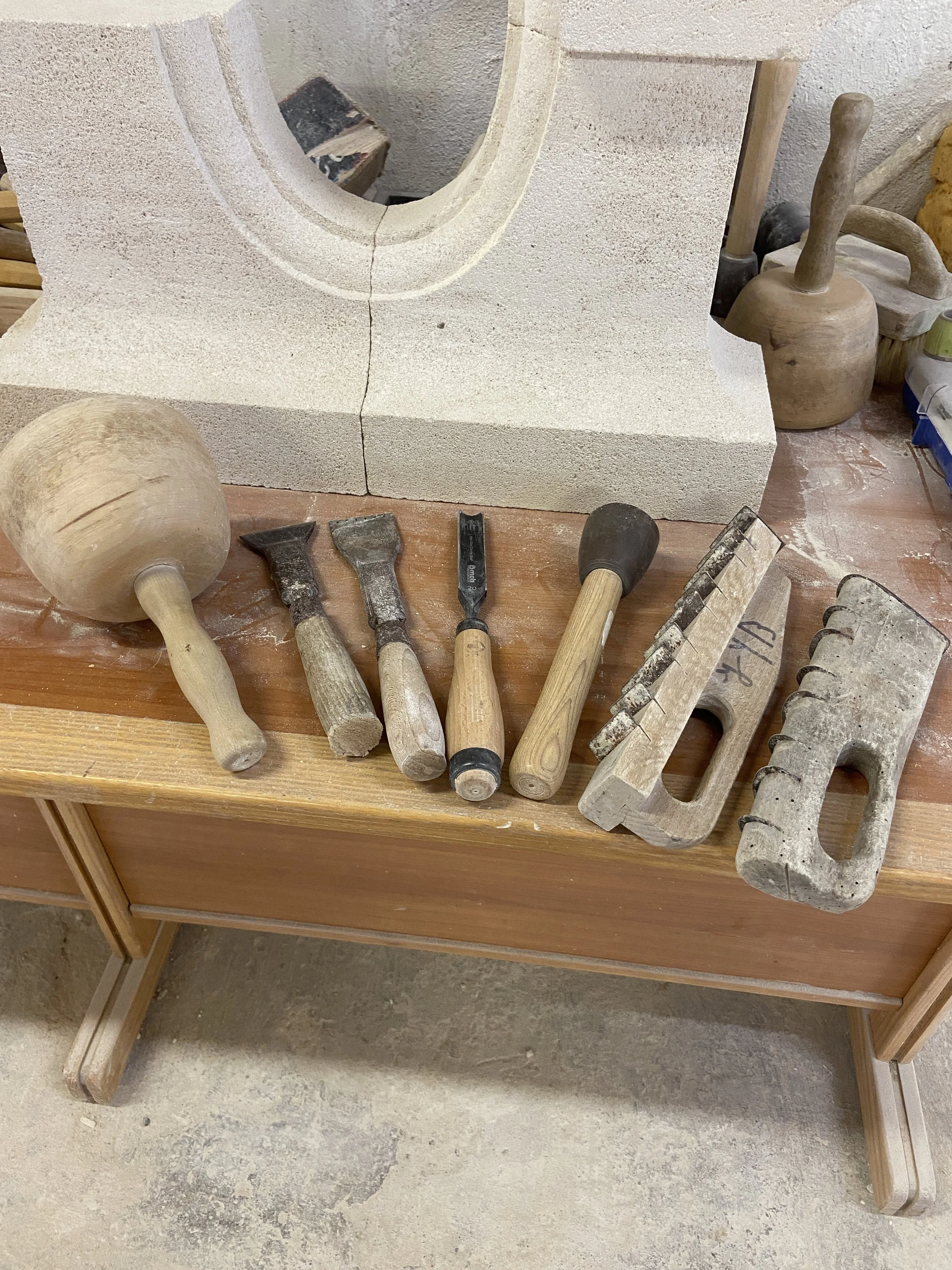 Outils de sculpture ou de céramique, y compris un marteau en bois, des gouges, un rasoir de sculpture, un maillet, une truelle, une spatule, et une forme en argile, organisés sur un plan de travail en bois avec de la poussière de pierre ou de terre autour.
