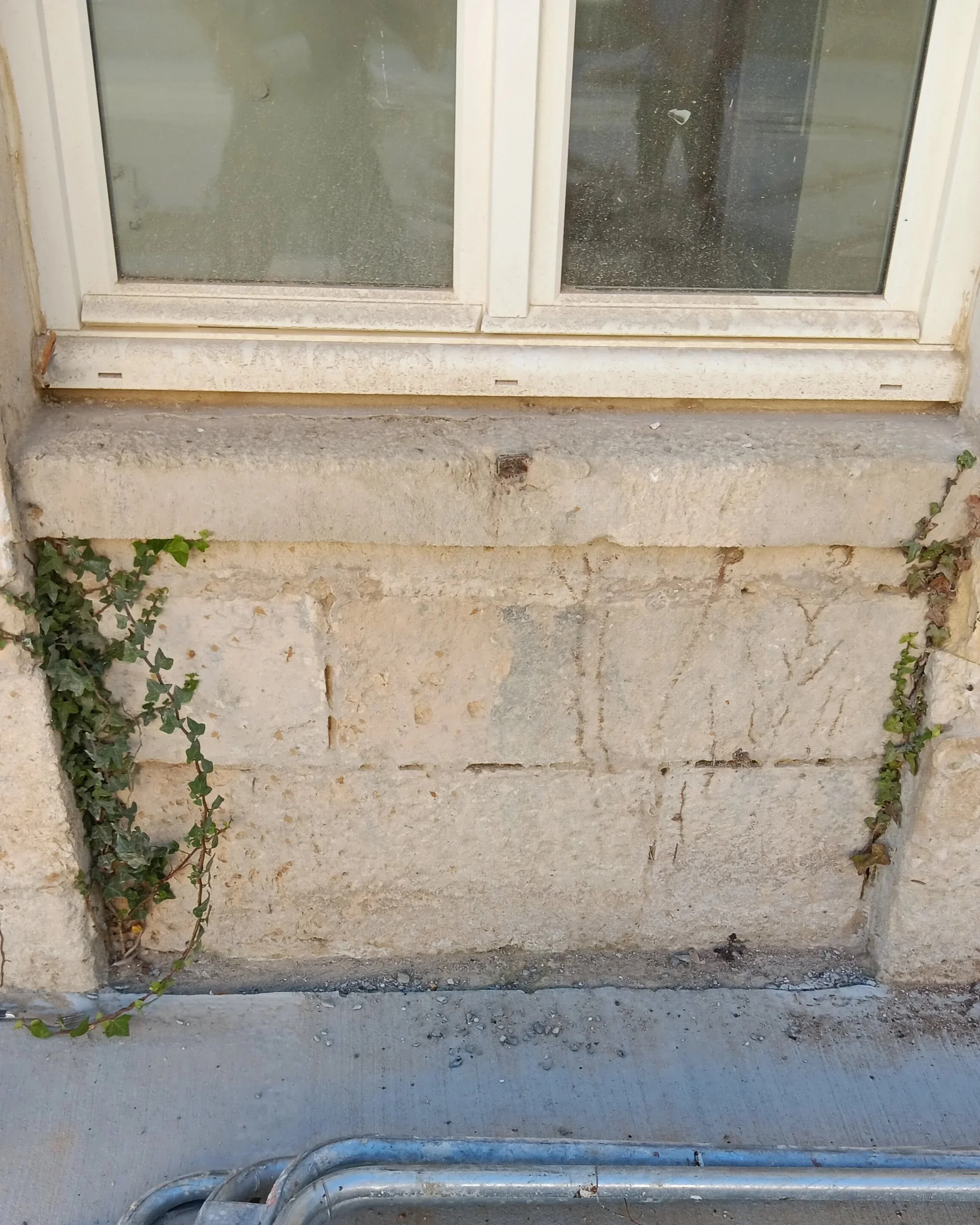 Fenêtre ancienne avec cadre en PVC blanc, sale et poussiéreux, installé sur un mur en pierre usée, avec des plantes grimpantes à gauche.