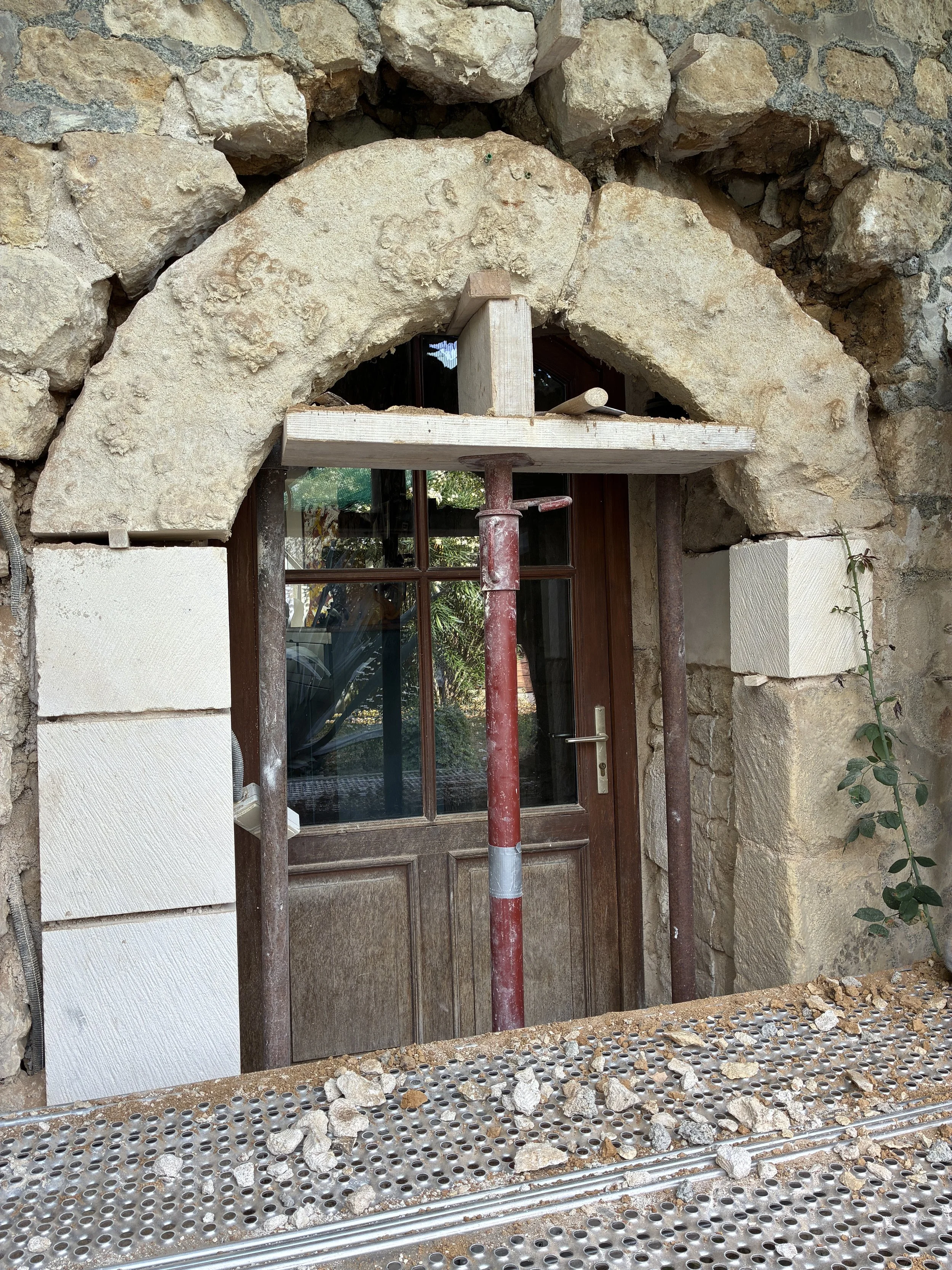 Construction en cours autour d'une porte en bois dans une façade en pierre, avec un échafaudage en métal et des pierres tombées sur la plateforme de travail.