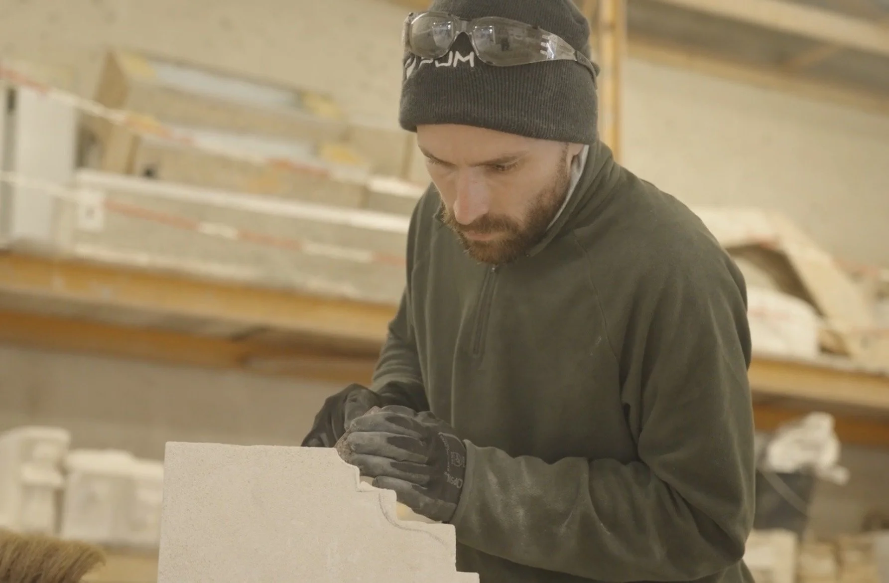 Un homme portant un bonnet, des lunettes de sécurité sur le bonnet, des gants, en train de travailler dans une usine ou une fabrication avec des matériaux blancs ou en pierre.