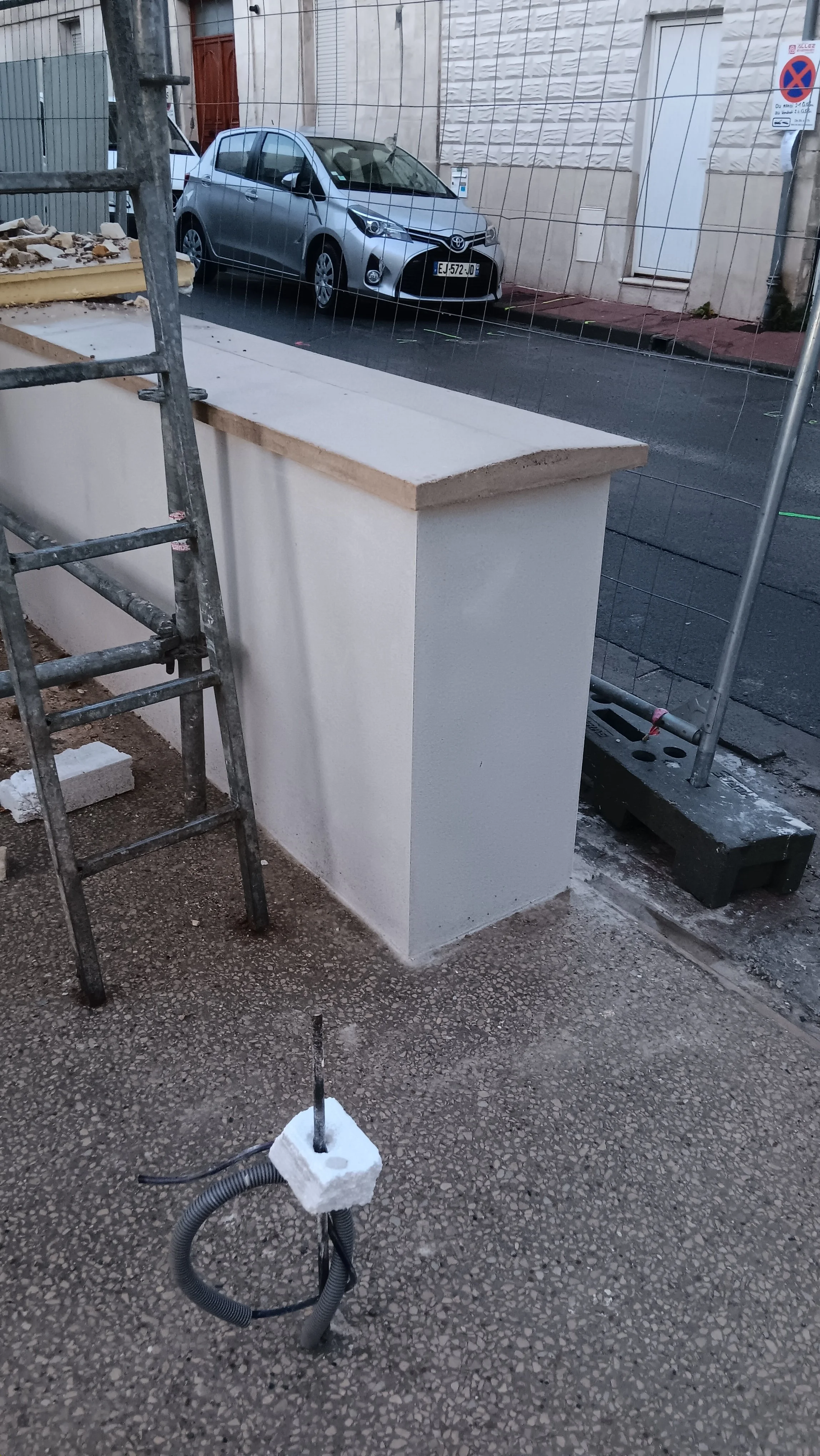 Construction ou rénovation en cours avec une échelle, un mur blanc, un poteau, et un câble électrique à terre, vue depuis l'intérieur vers la rue avec une voiture garée dehors.