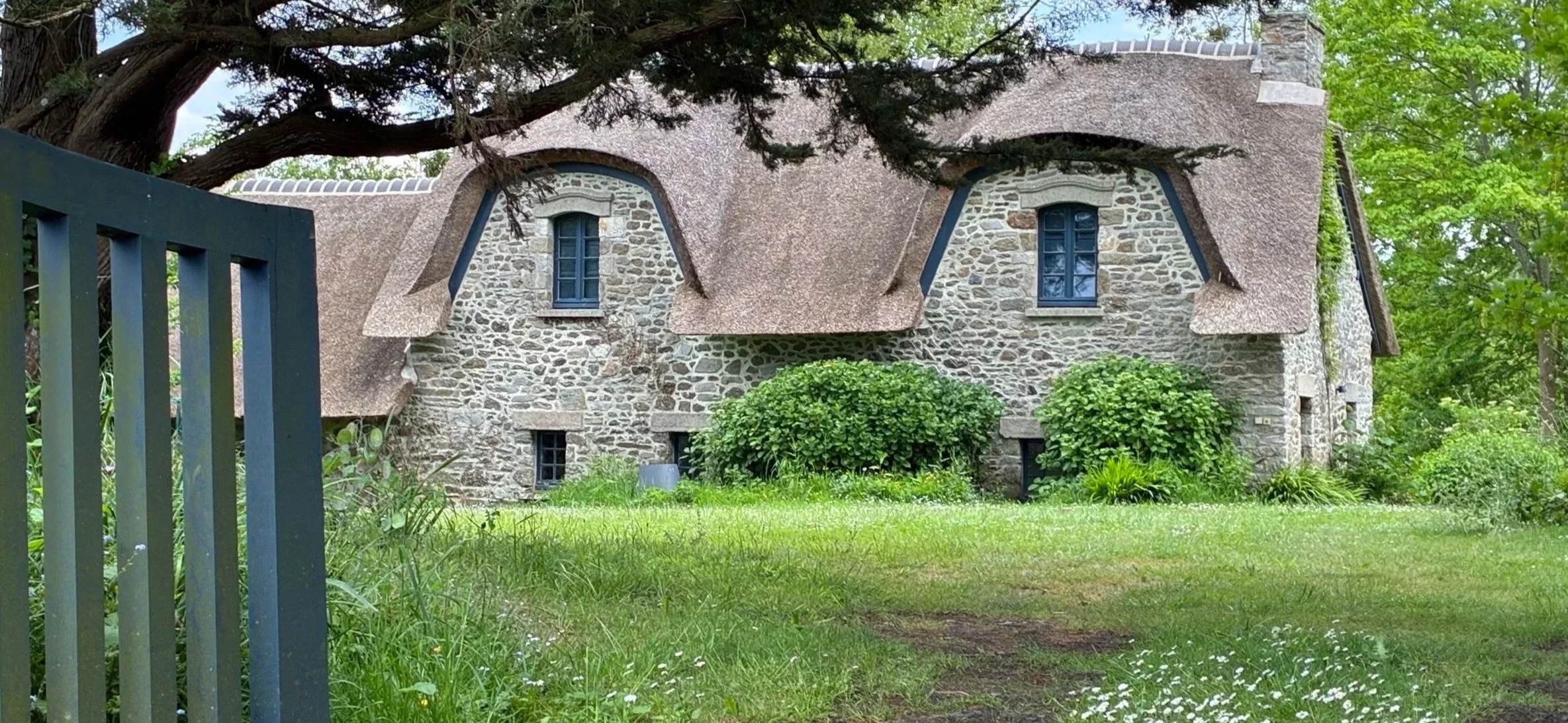 Maison en pierre avec toit en chaume, entourée d'arbres et de végétation verdoyante.
