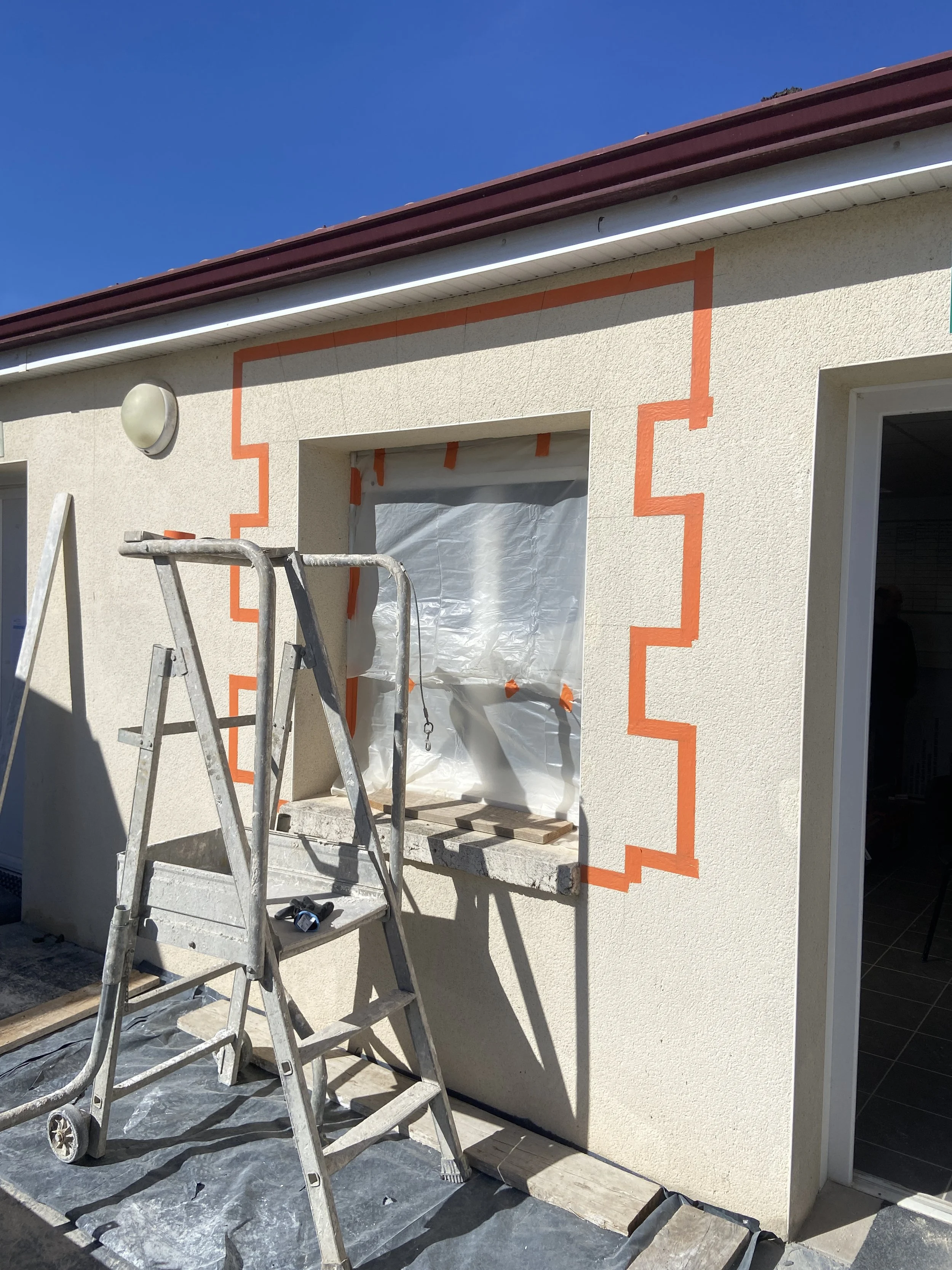 Travaux de peinture en cours sur une façade de maison avec des bandes de protection orange autour de la fenêtre, une échelle en aluminium devant la façade, un plafonnier rond et une porte ouverte.