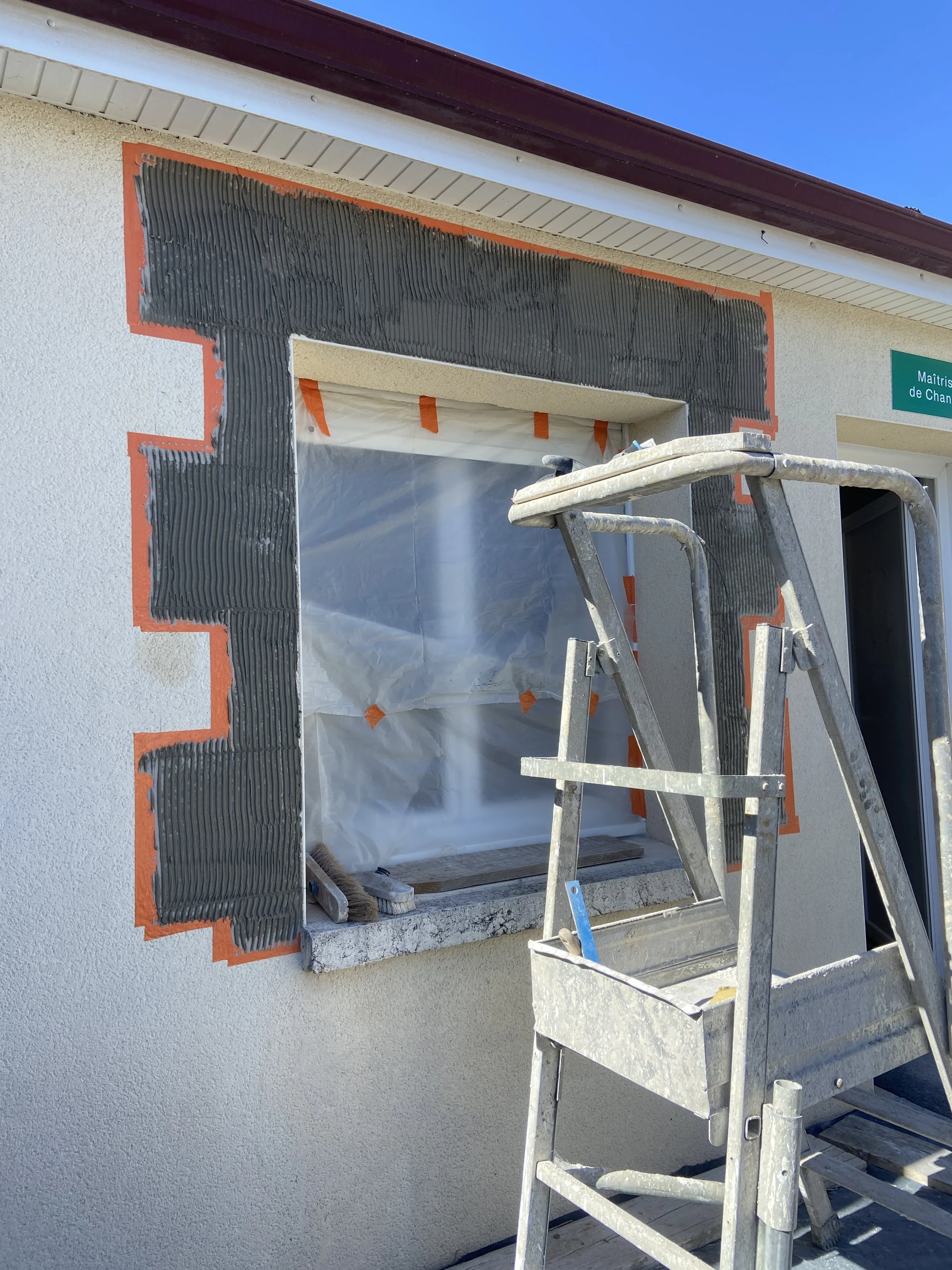 Construction d'une fenêtre encadrée en noir sur une façade de maison, avec un échafaudage en aluminium devant la fenêtre. La maison a une façade beige et un garde-corps blanc, un toit en tuiles marron, et le ciel est bleu.
